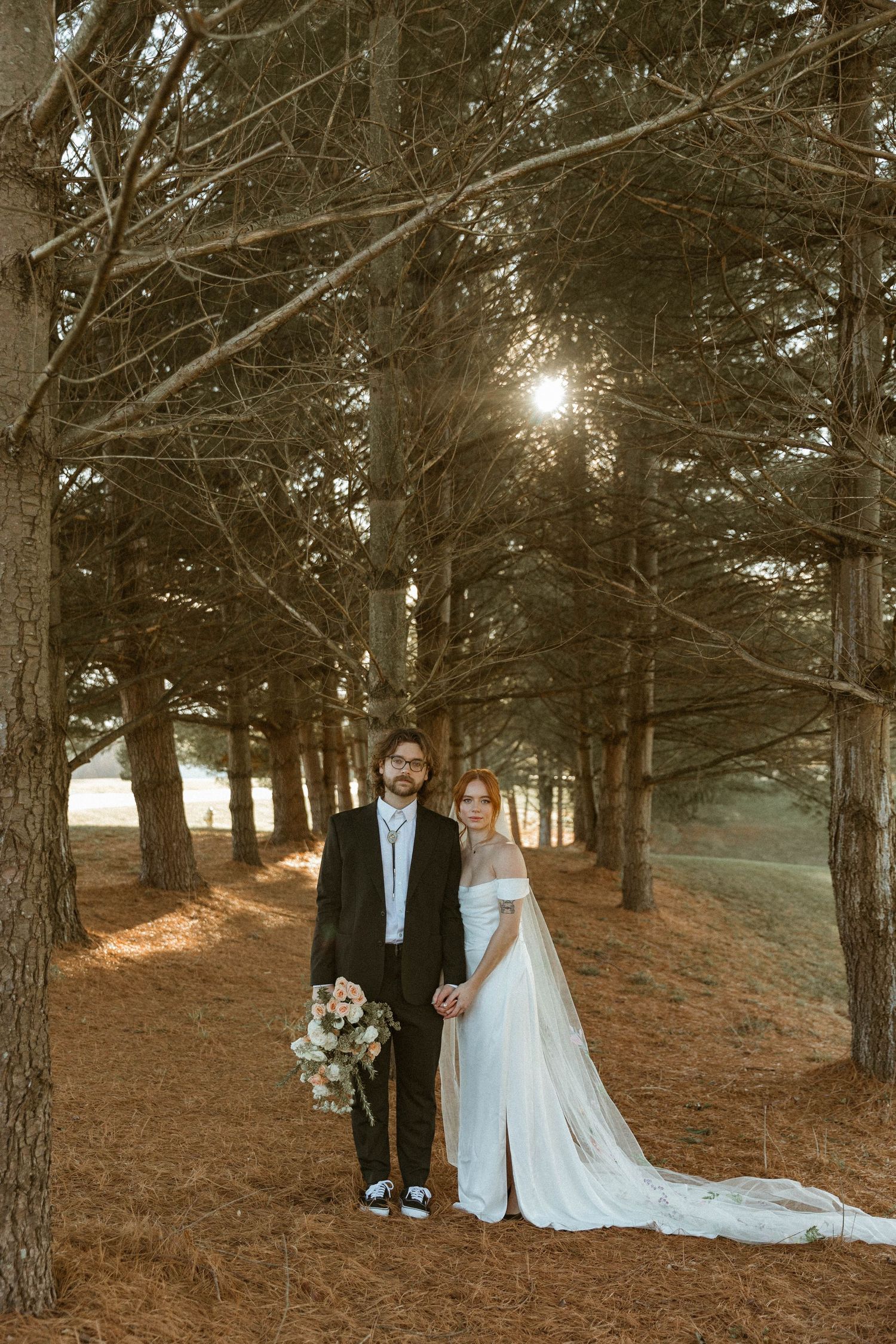 Bride and groom posing together among pine trees at sunset in a romantic forest setting.