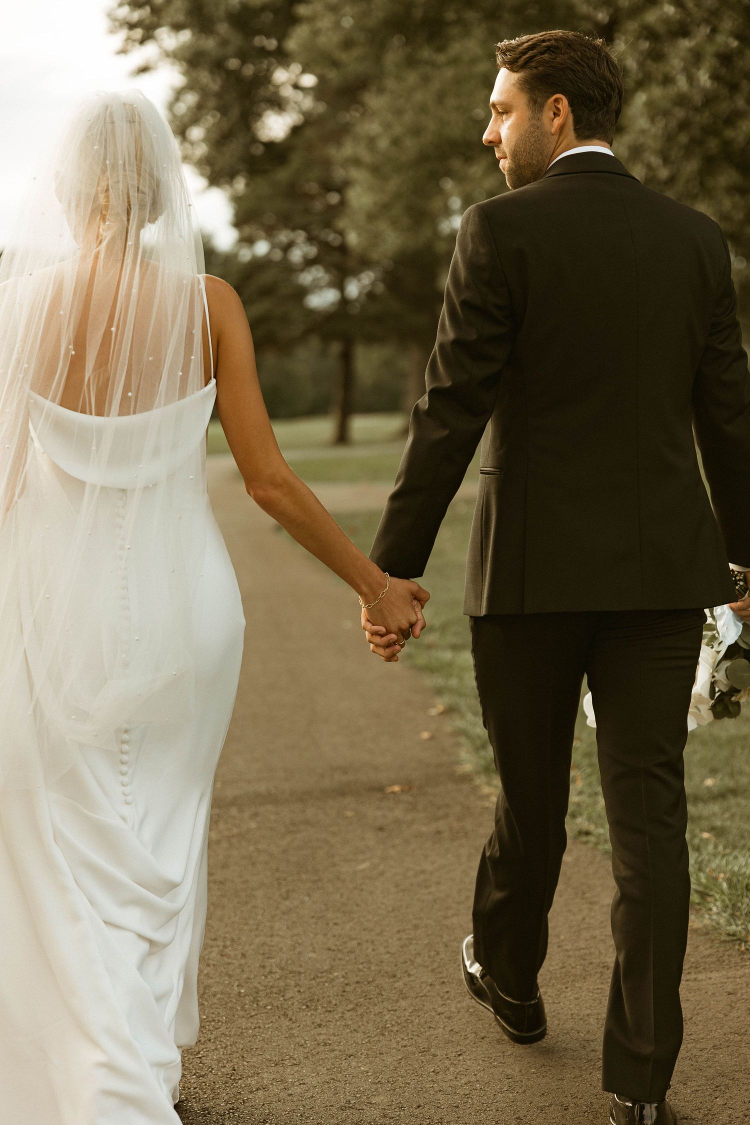 Couple holding hands walking down park path in elegant wedding attire.