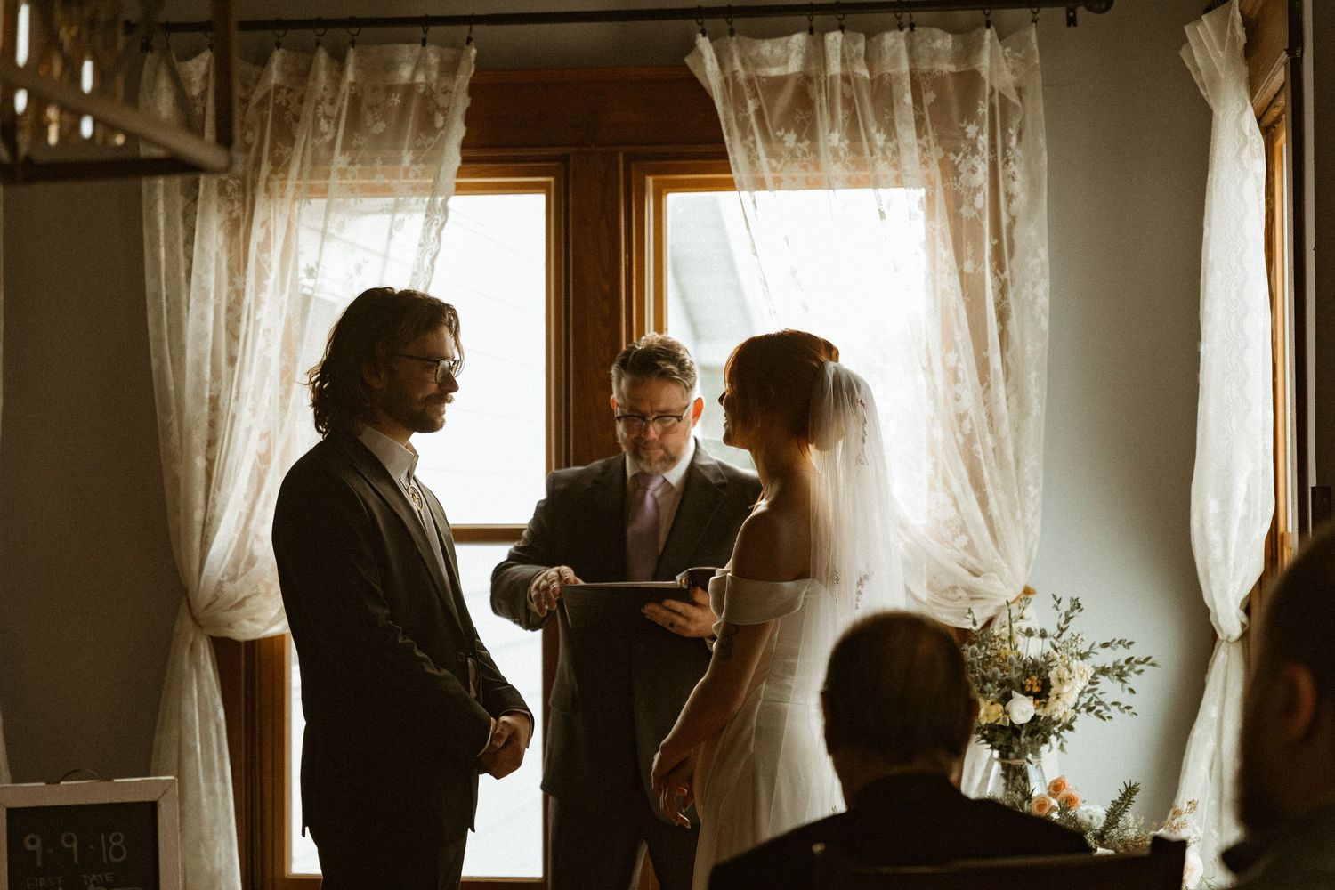 Wedding ceremony in progress inside a rustic room with lace curtains and natural lighting.