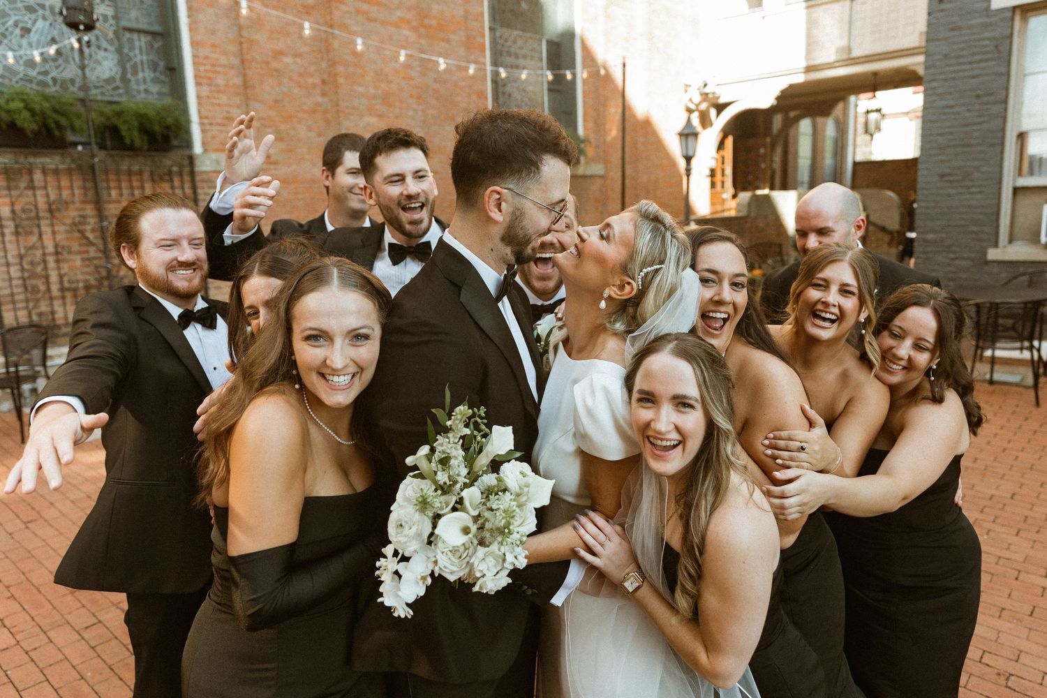 Wedding party members embracing and laughing together outside brick building.