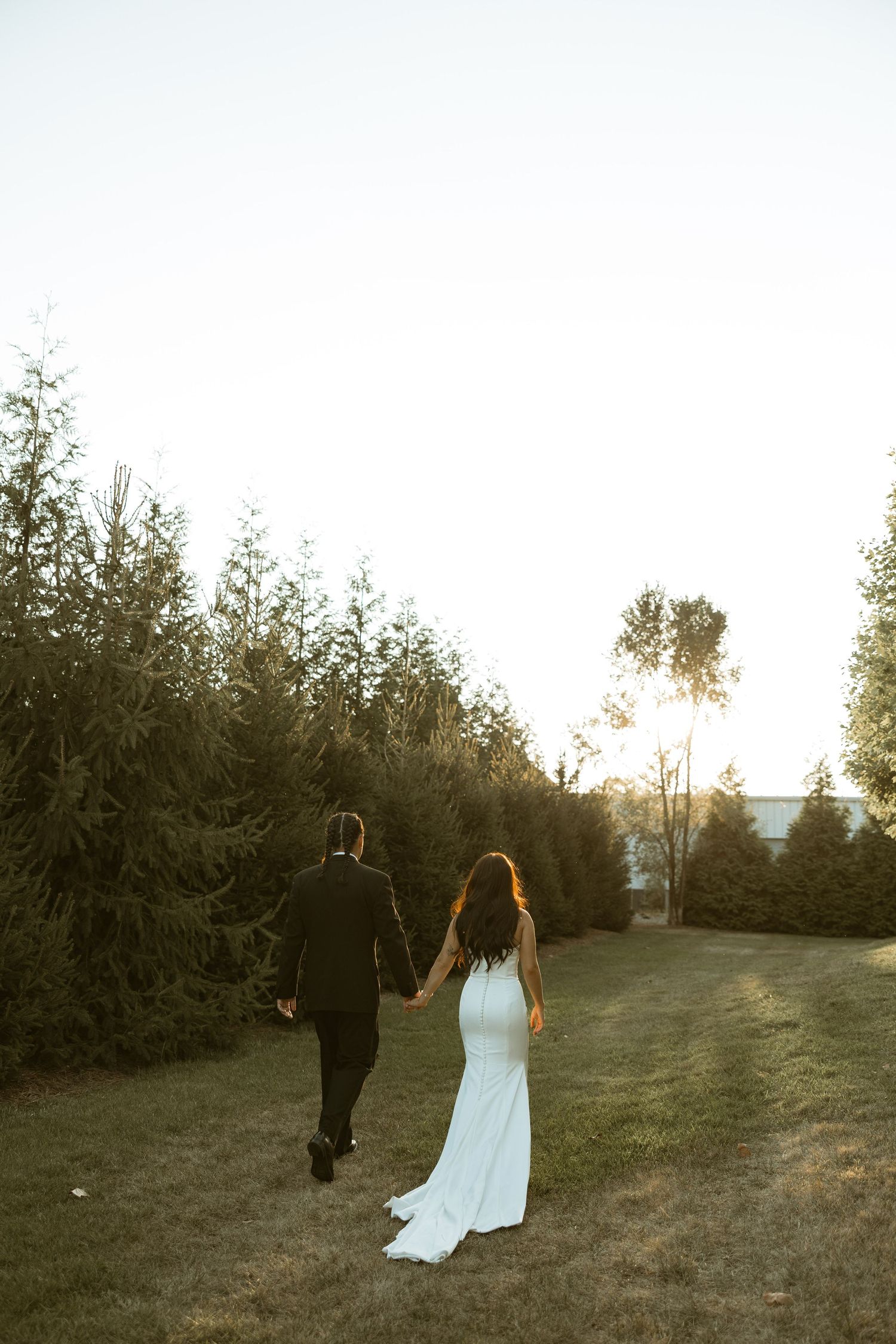 A couple walking hand in hand on a grassy path at sunset with trees in the background.