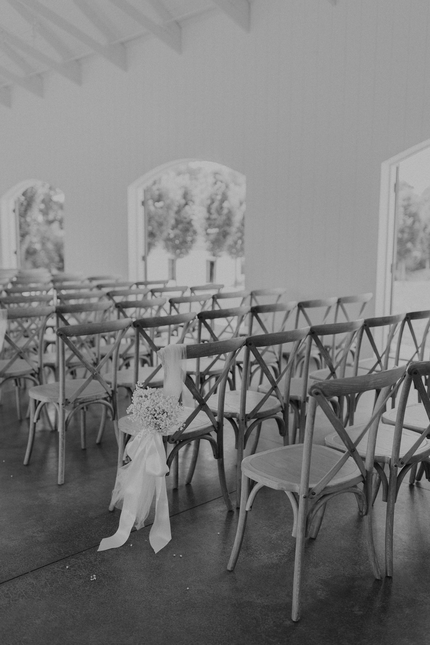 Empty rustic wooden chairs arranged for a wedding ceremony in a white room with arched windows.