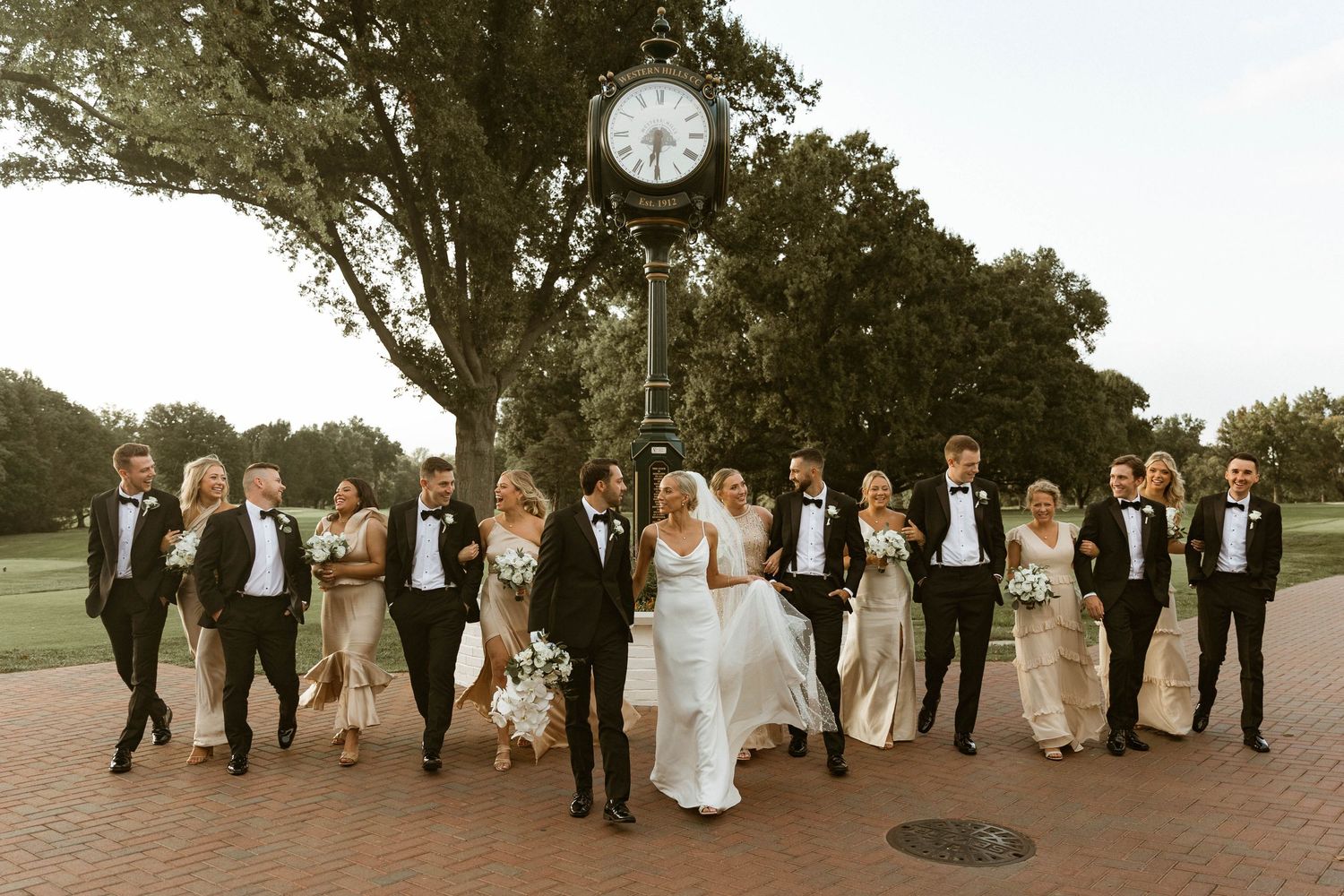 Wedding party celebrating together under trees near vintage clock on campus grounds.