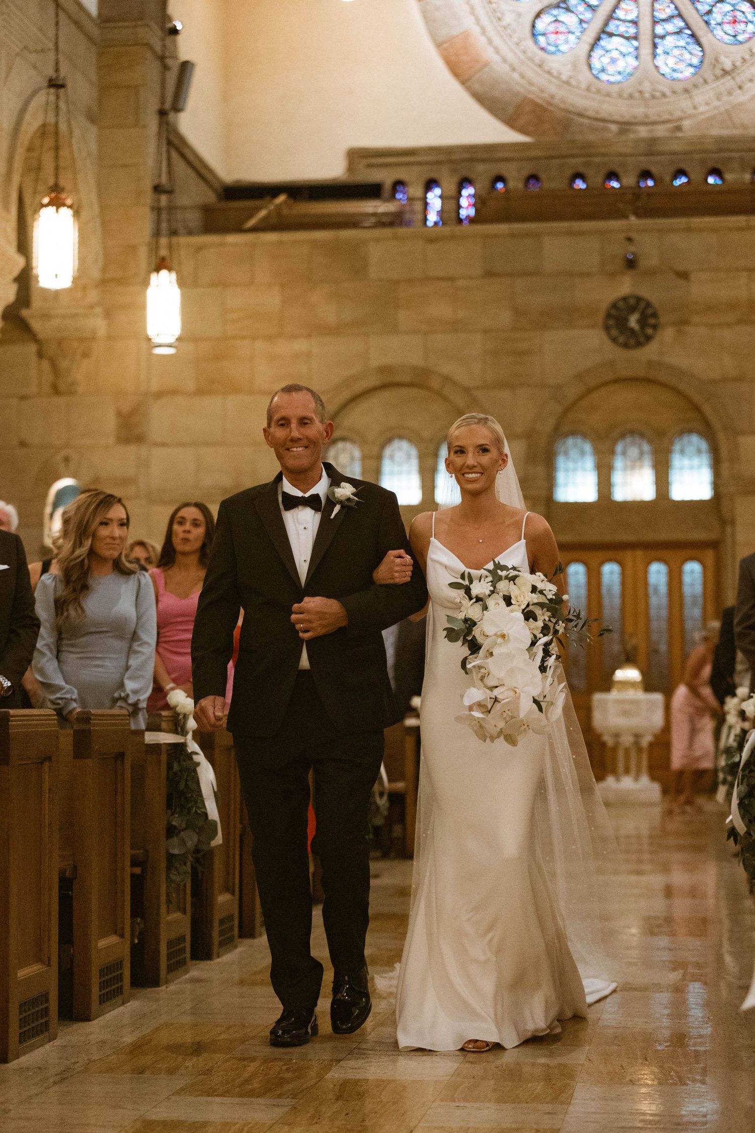 Wedding ceremony procession in ornate church sanctuary with stained glass windows.