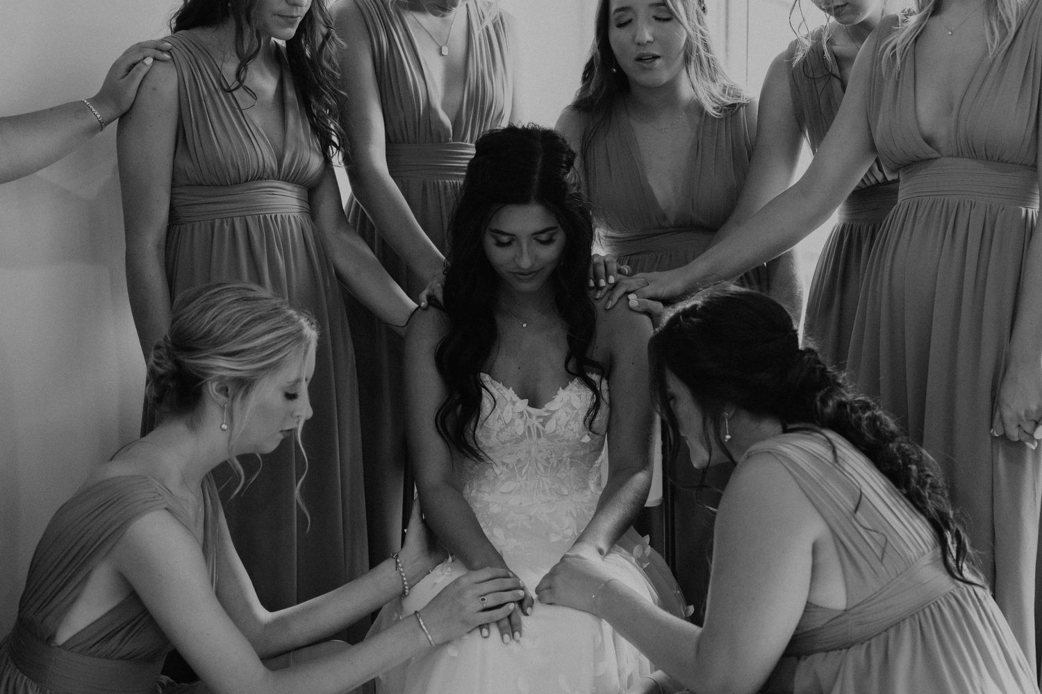 Bridesmaids in pale dresses gather around a seated bride in a white lace gown for a special pre-wedding moment.