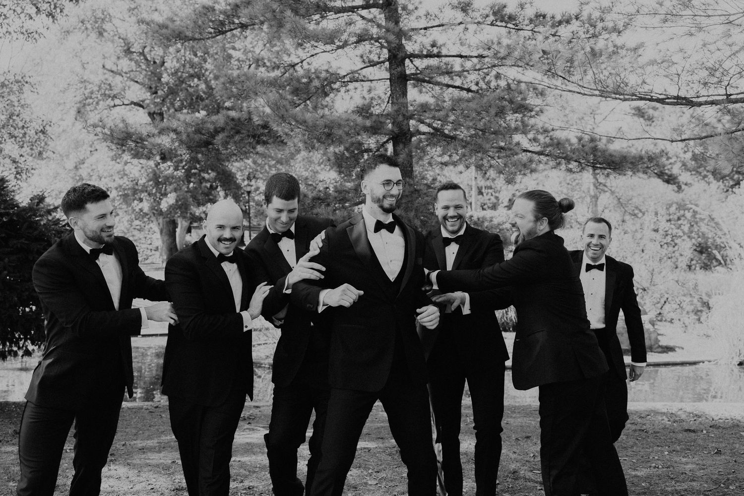 Groomsmen in black tuxedos posing for fun group photos outdoors.