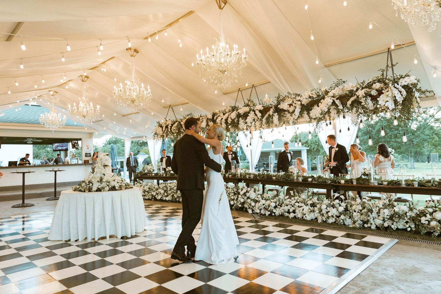 Newlyweds share their first dance under crystal chandeliers on a checkered dance floor.