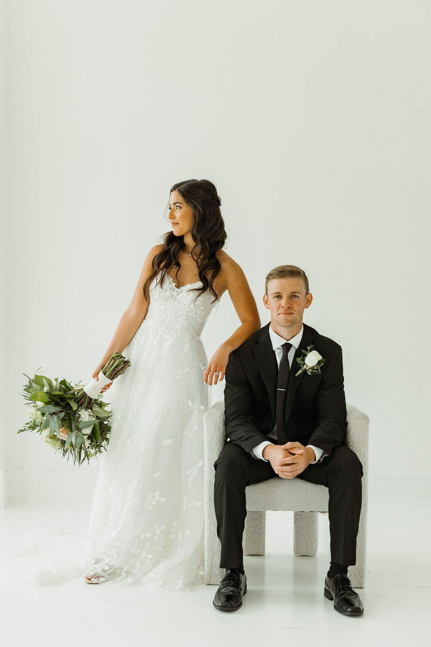 A stylish wedding couple poses in studio against white backdrop with the bride holding a bouquet.
