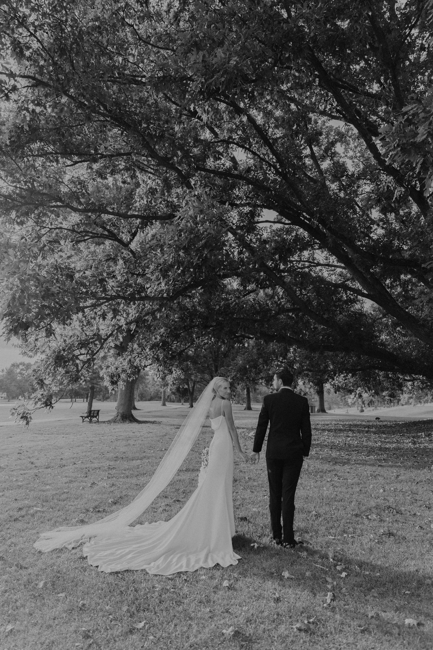 Black and white photo of newlyweds walking under large trees with flowing veil.
