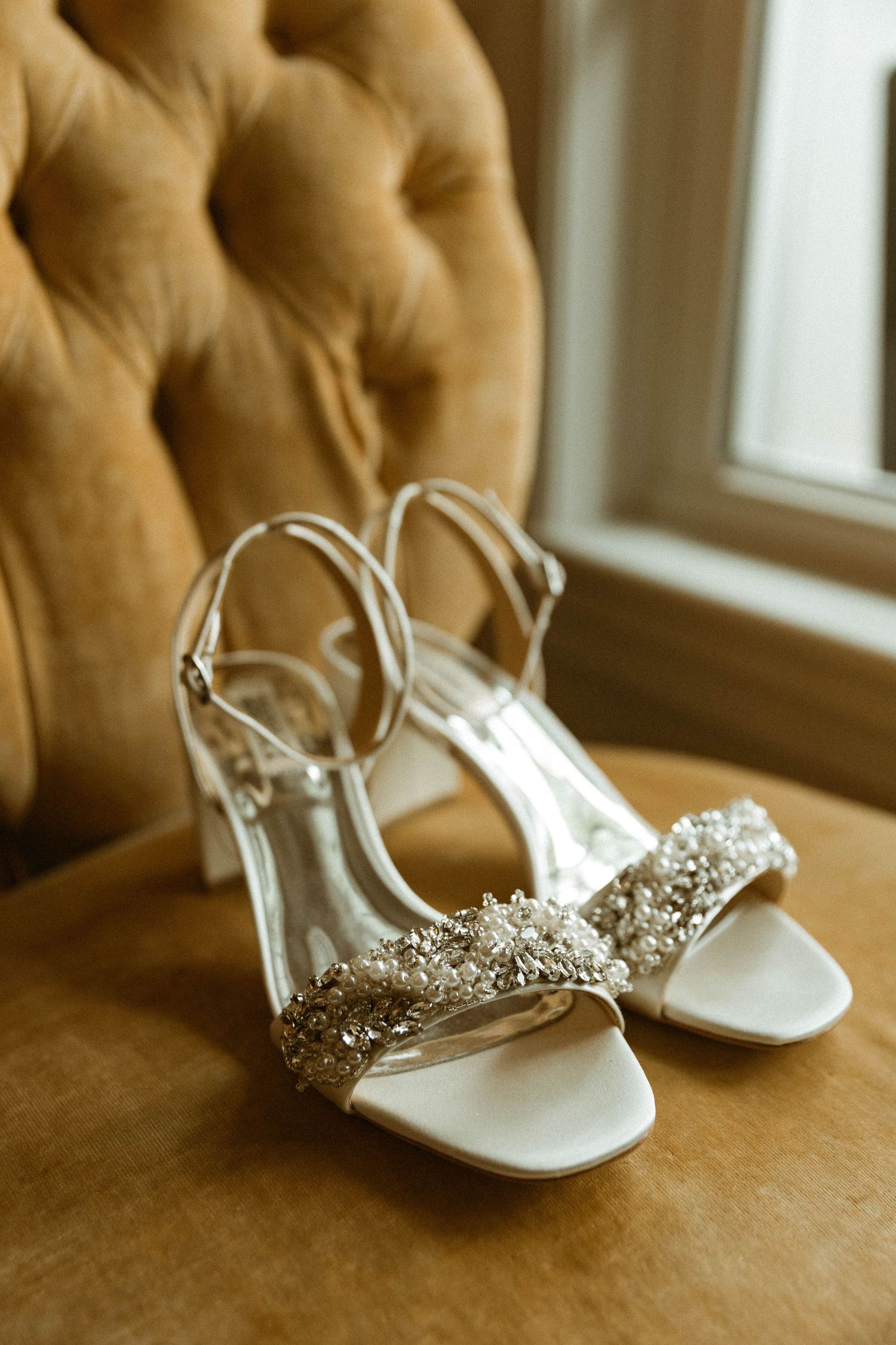 Crystal-embellished white bridal sandals on a tufted tan leather chair.