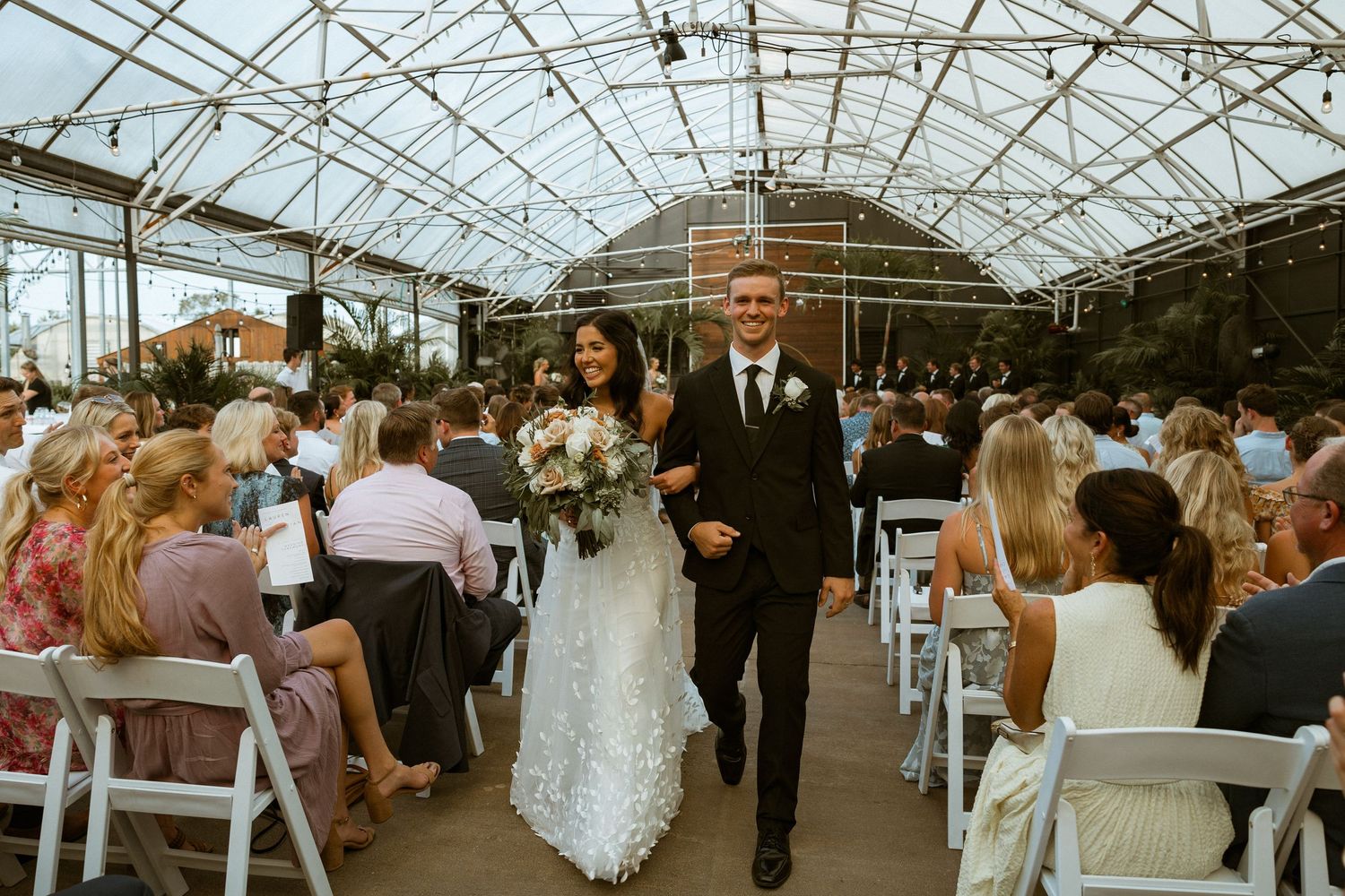 Wedding ceremony and reception setup inside a modern greenhouse with glass ceiling.