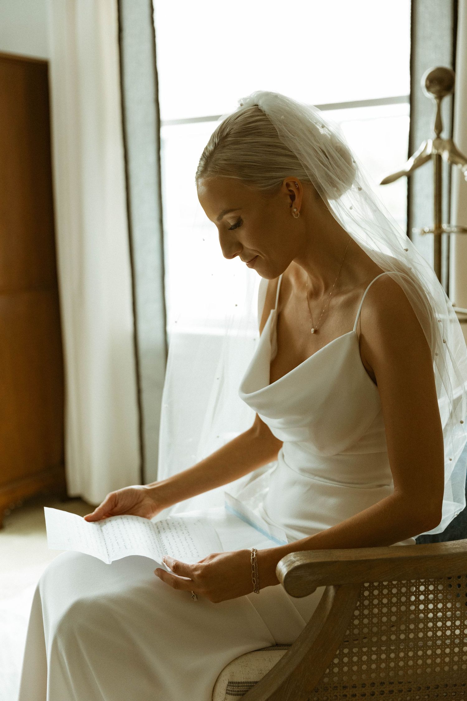 Bride in elegant white gown reading document while seated by window.