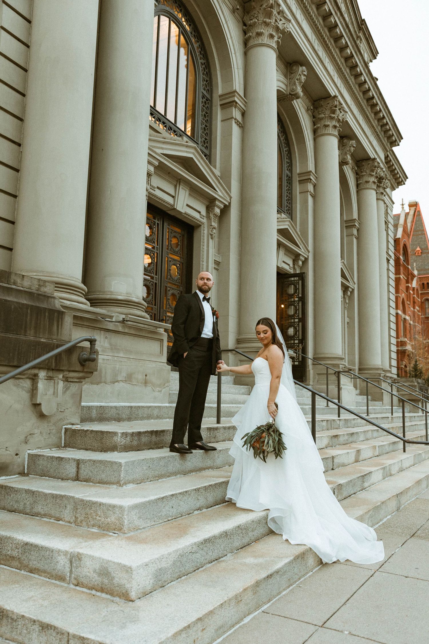 A wedding couple poses on grand stone steps of a historic building with majestic columns.