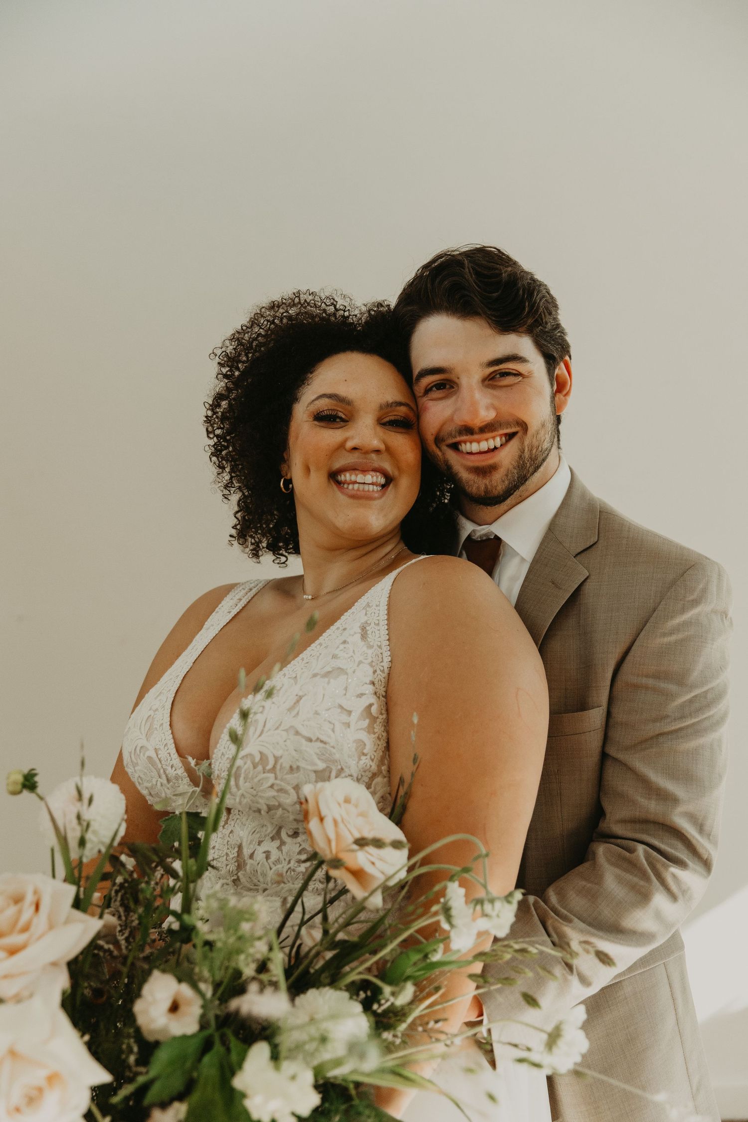 Joyful wedding portraits of couple with floral bouquet against neutral background.