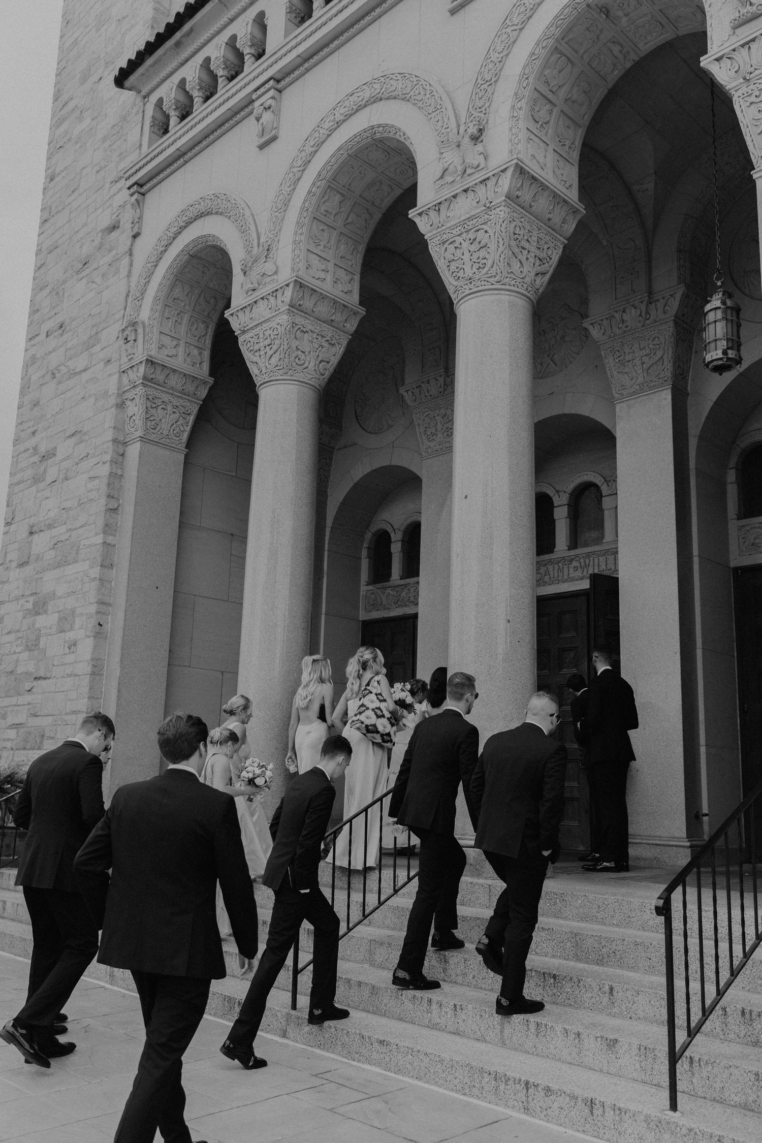 Wedding party ascending church steps under ornate stone arches.