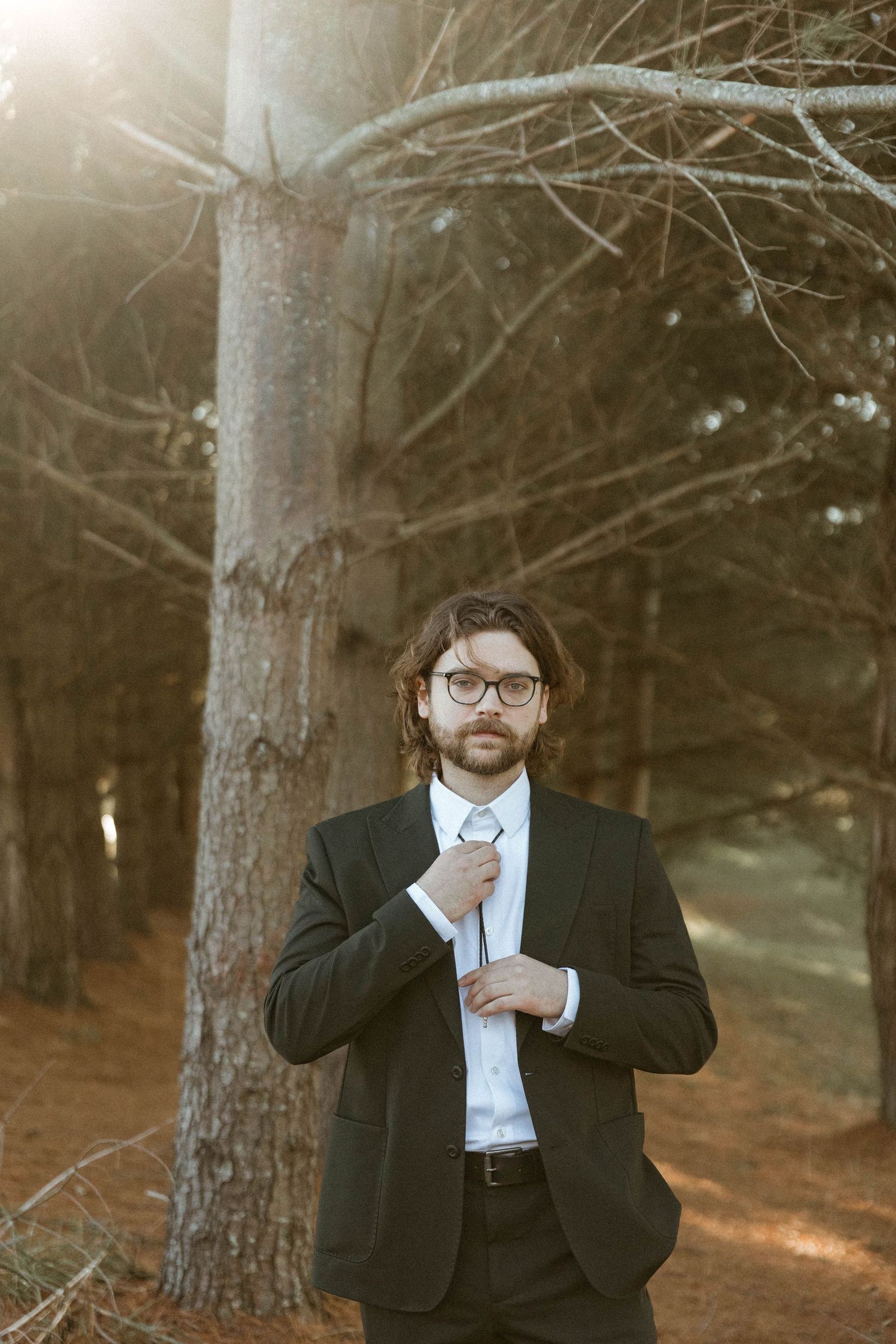 A suited figure adjusts their tie while standing among trees in golden afternoon light.