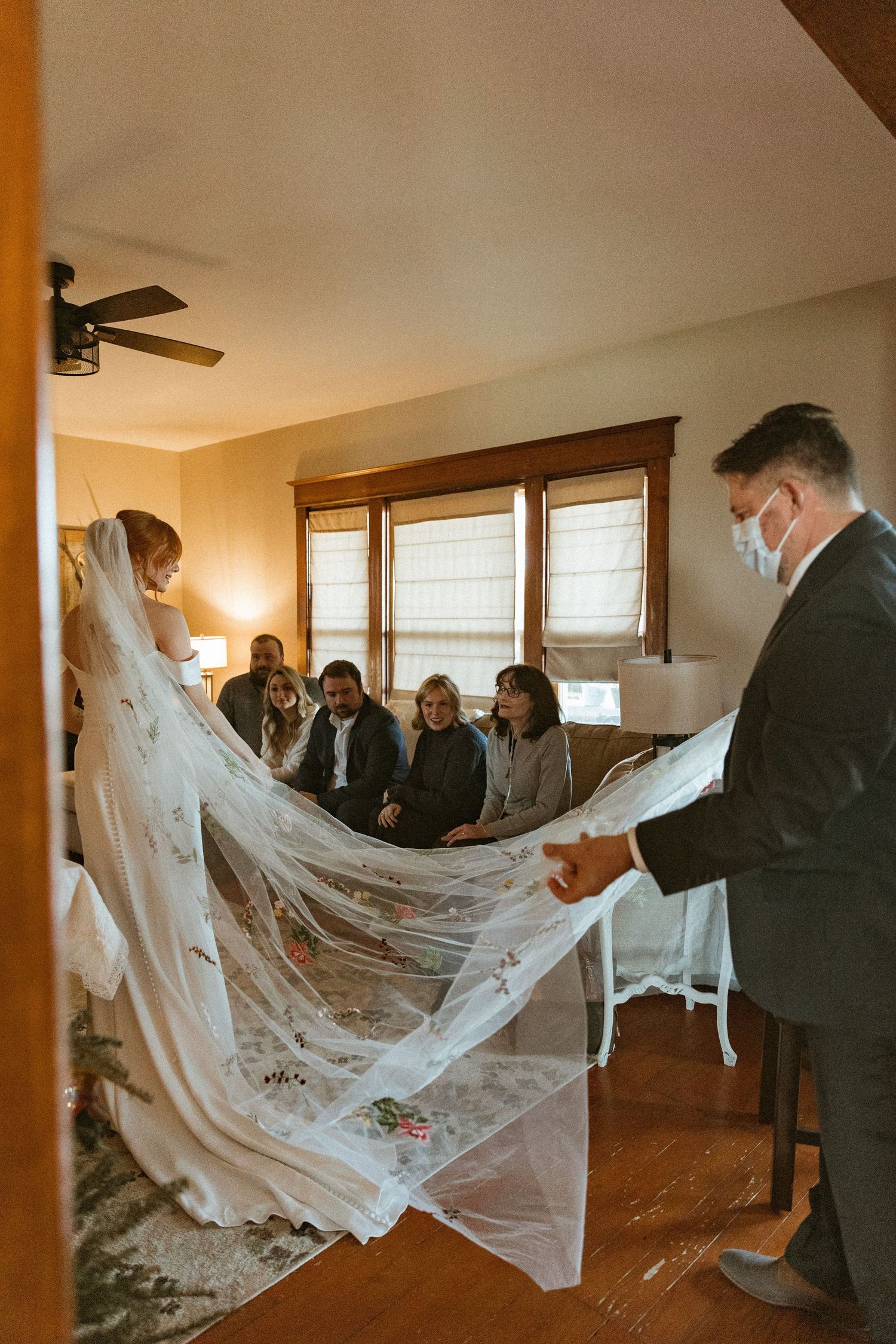 Wedding preparations showing attendants helping with the bride's long veil in a sunlit room.