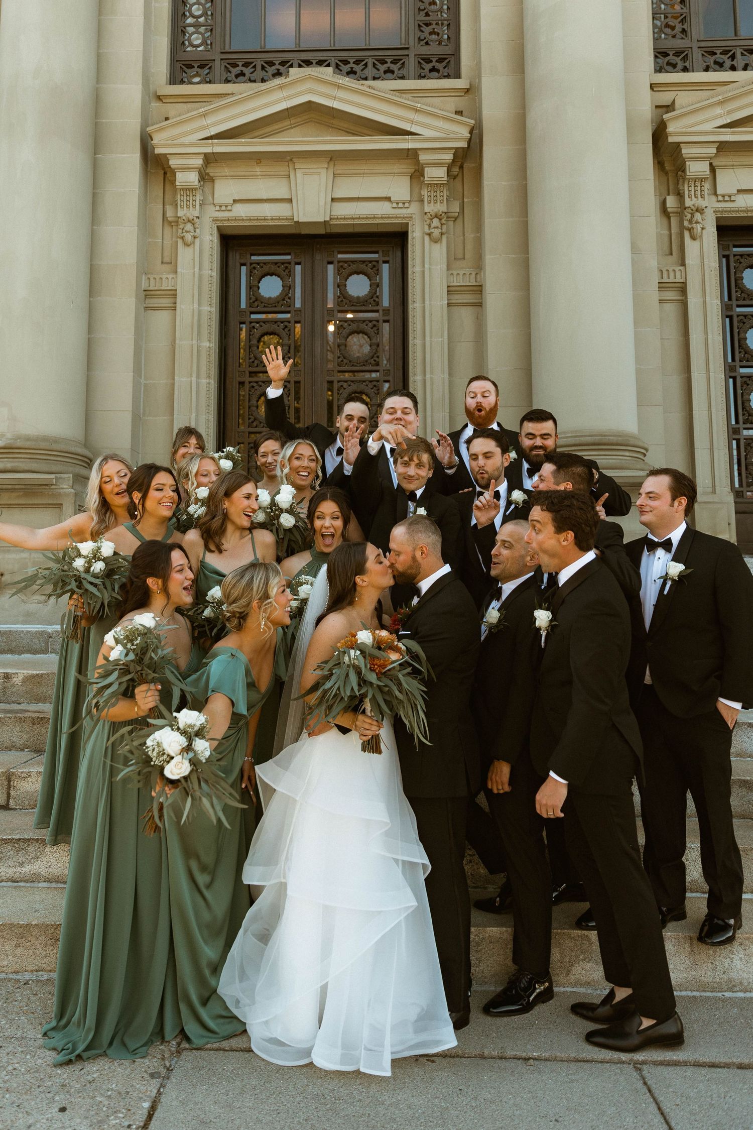 Wedding party photo on stone steps with bridesmaids in sage dresses and groomsmen in black tuxedos.