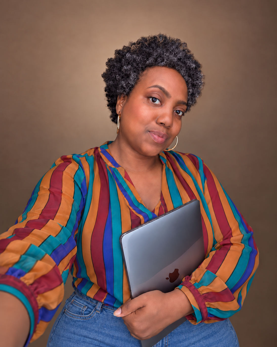 Woman in colorful striped blouse holding a laptop, wearing hoop earrings, posing confidently against a brown background.