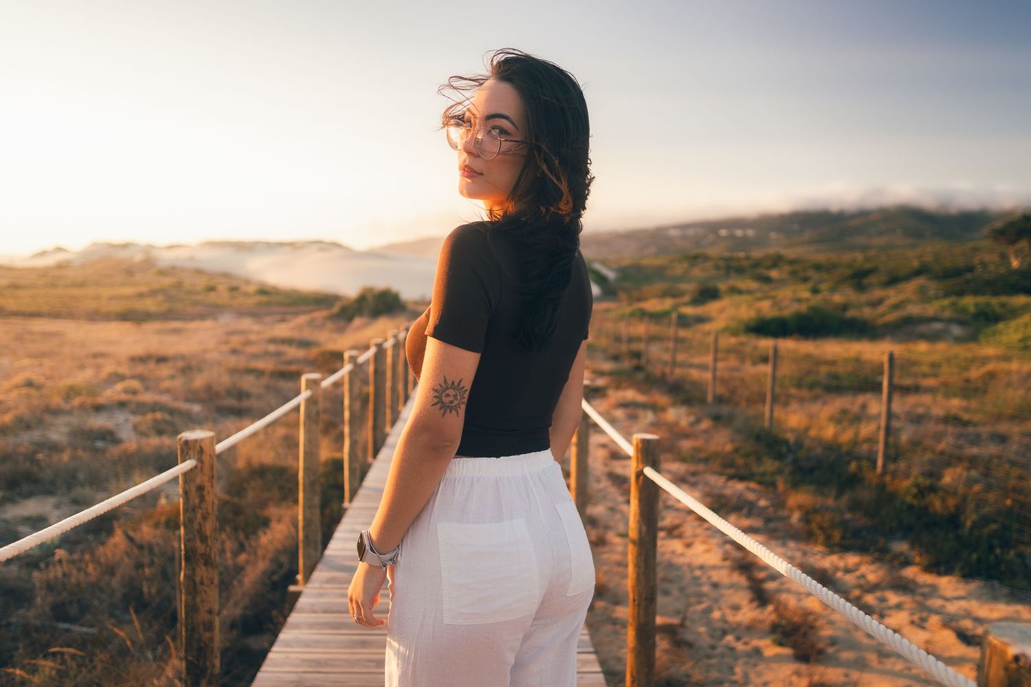 A figure stands on a wooden boardwalk path at sunset with golden light illuminating rolling hills in the background.