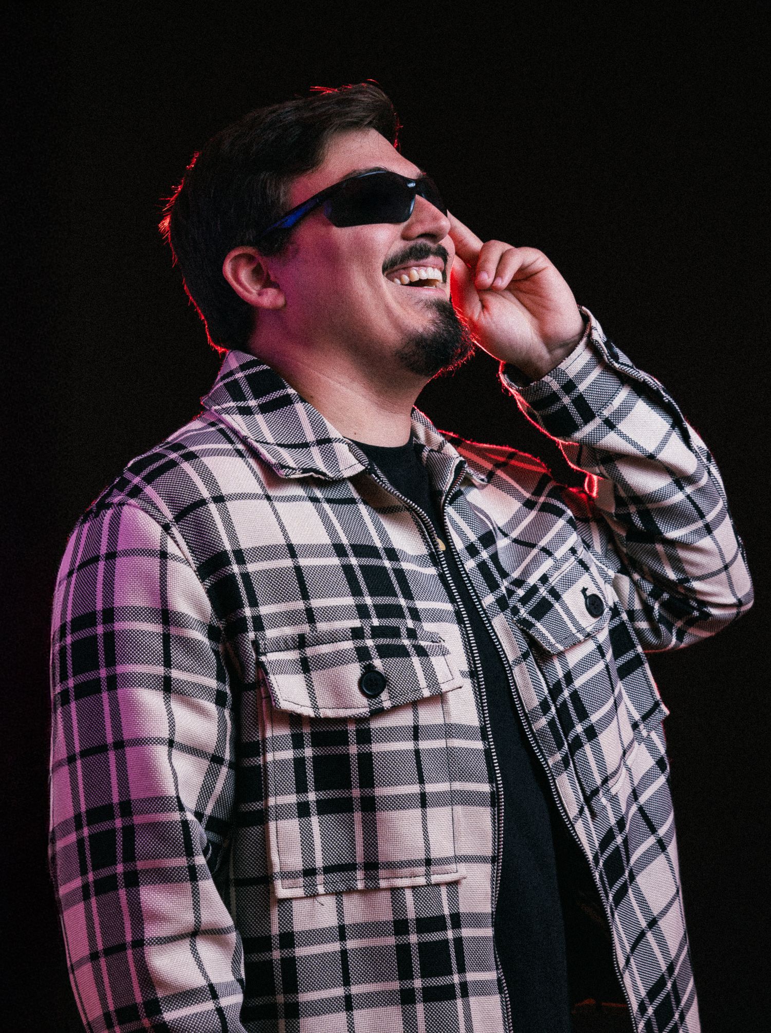 Performer in plaid shirt singing passionately on dark stage with red lighting effects.