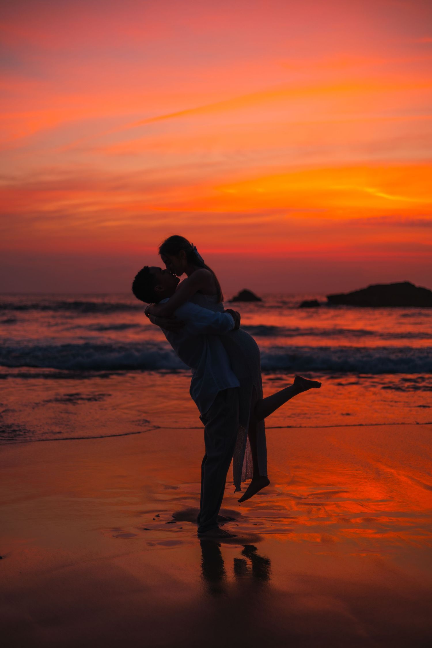 Silhouettes of couple sharing romantic moment against vibrant orange sunset on beach.