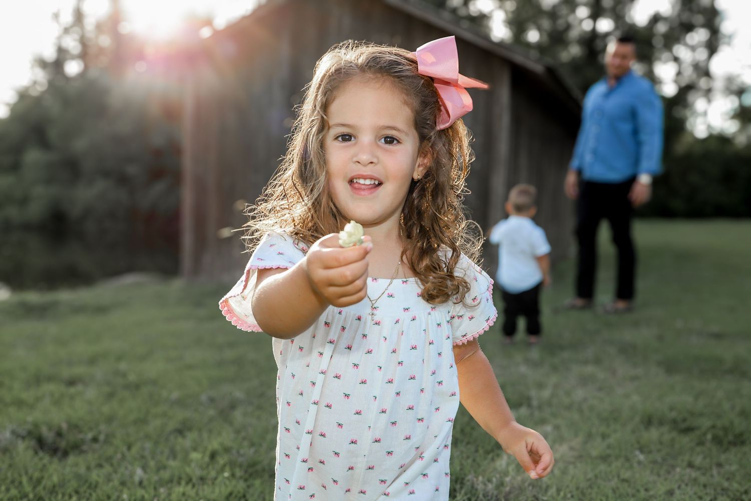 Child in white dress with pink bow reaching forward outdoors at sunset.