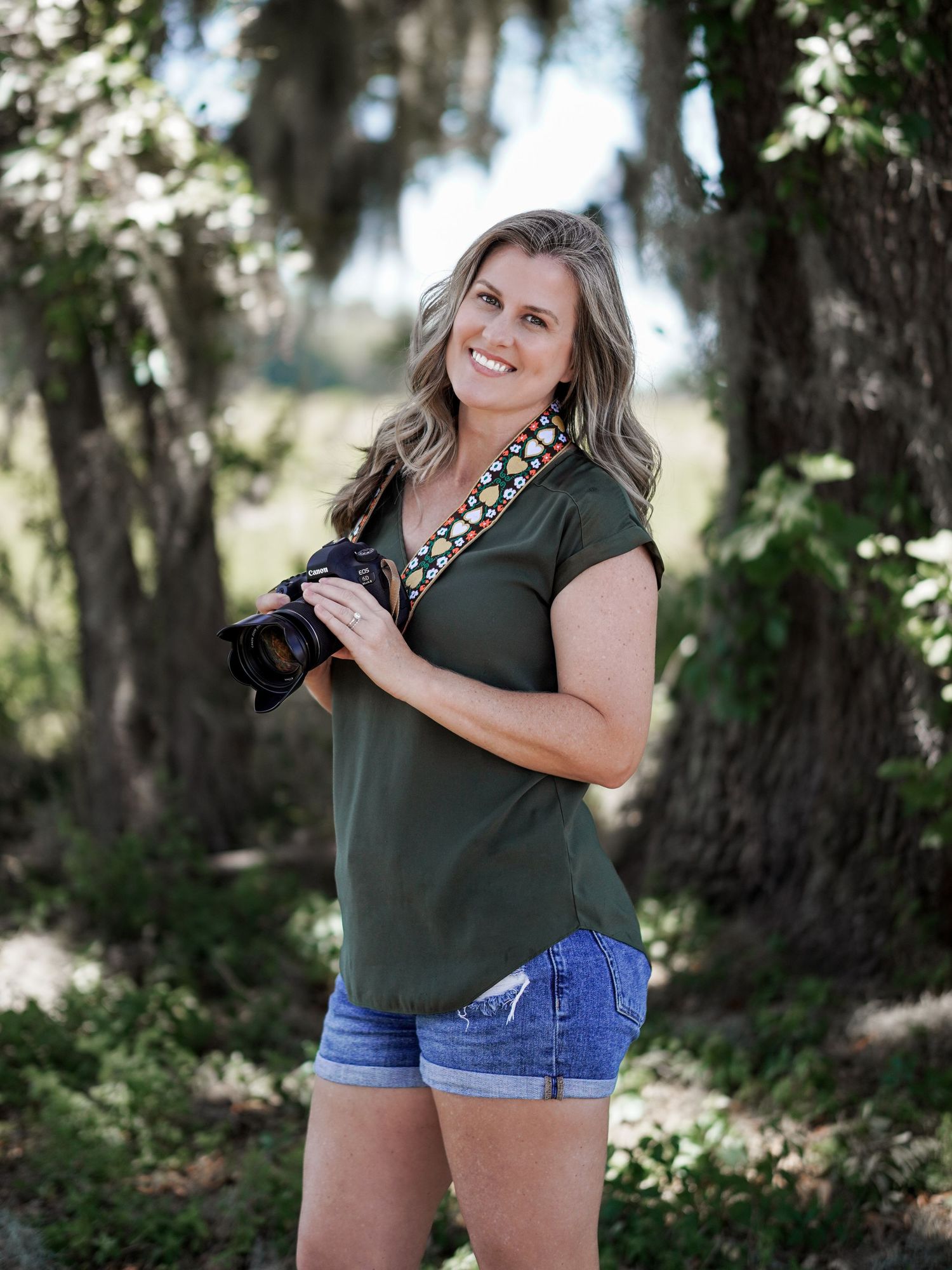 Woman in olive green shirt holding camera while standing among moss-draped trees.