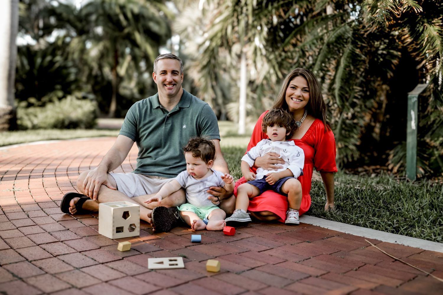 A family sits together playing with wooden toys on a brick pathway surrounded by lush palms.
