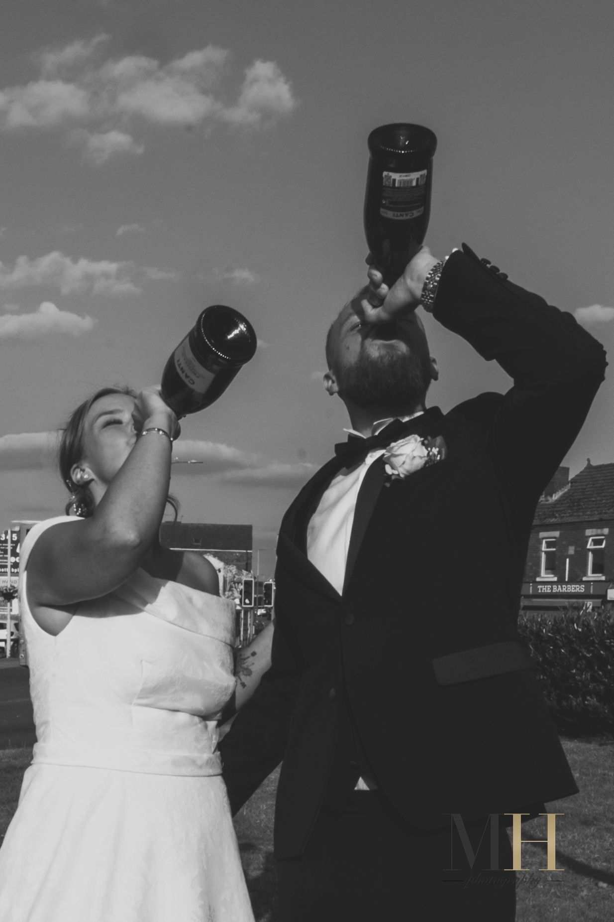 Wedding couple takes celebratory drinks together against bright sky background.