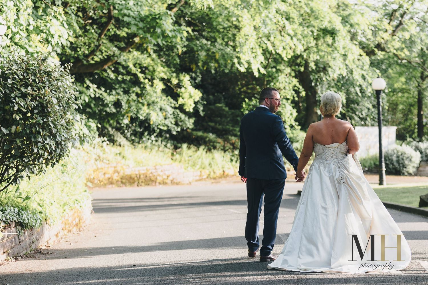 A bride and groom walk hand in hand down a tree-lined path on their wedding day.