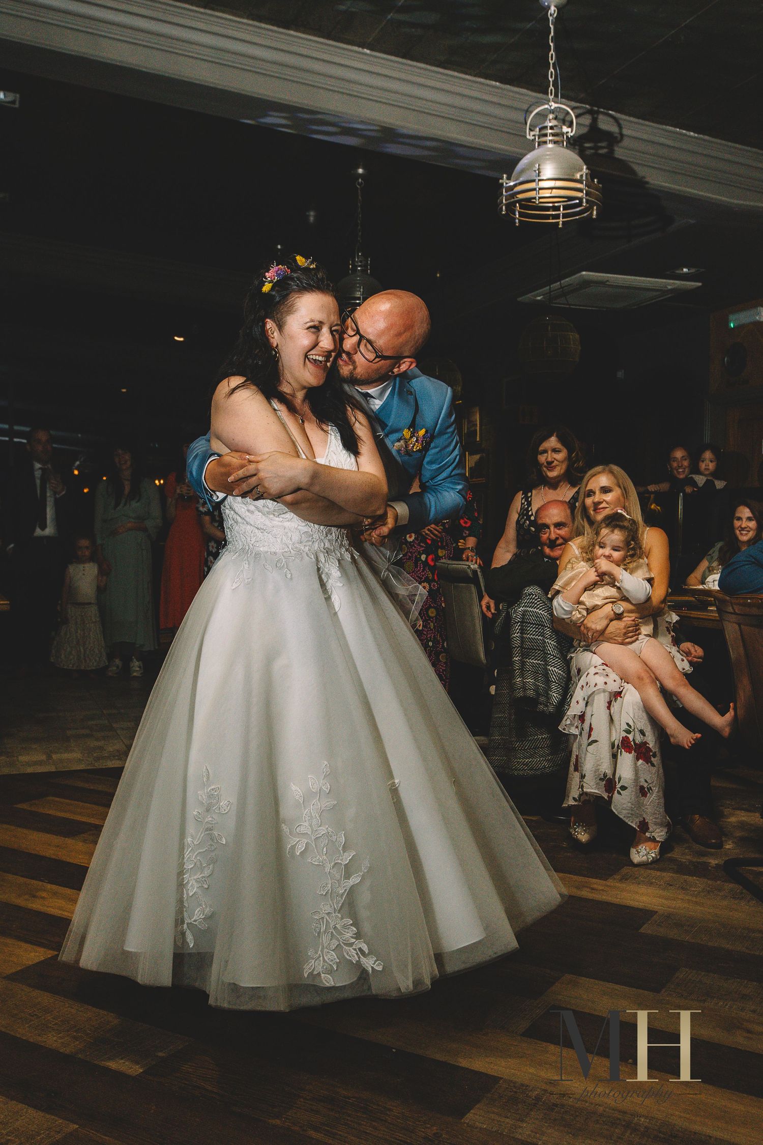 A couple shares their first dance at their wedding reception while guests watch from the sidelines on a wooden dance floor.