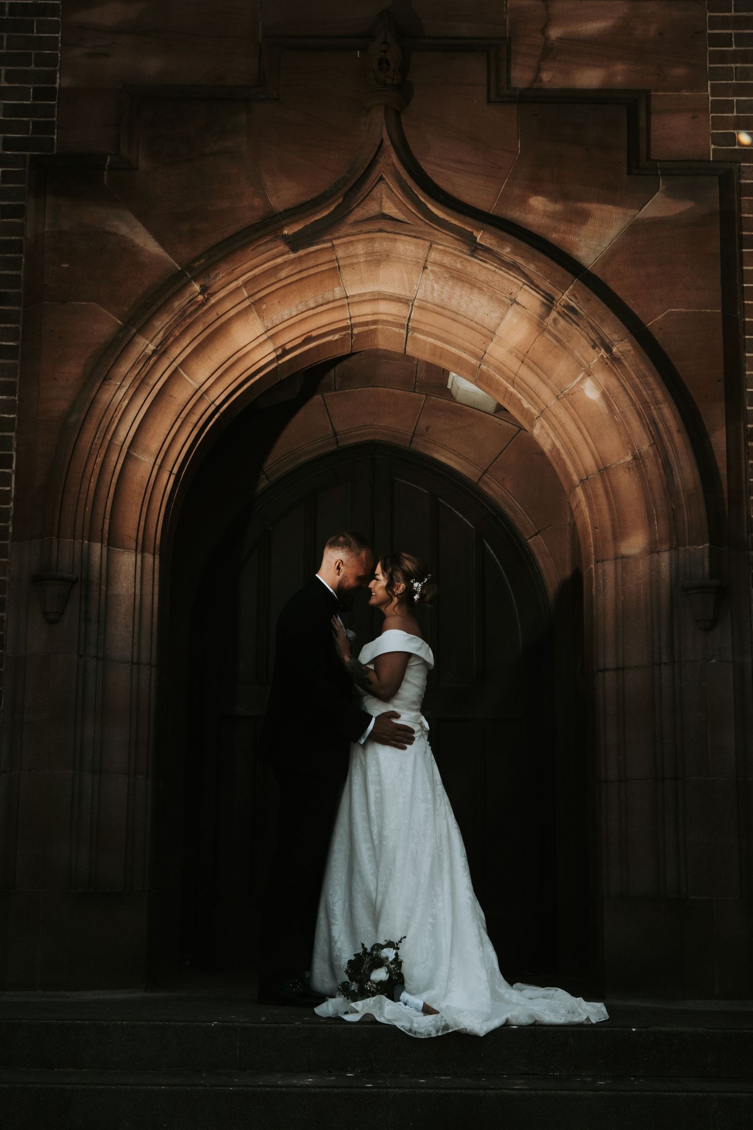 Romantic wedding couple embraces in front of gothic stone archway with dramatic lighting and flowing white dress.