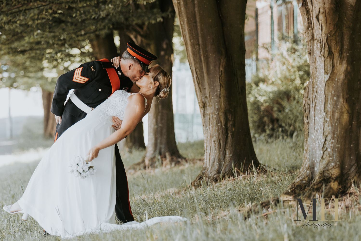 Military groom in dress uniform dips bride in white wedding gown for romantic kiss outdoors among trees.