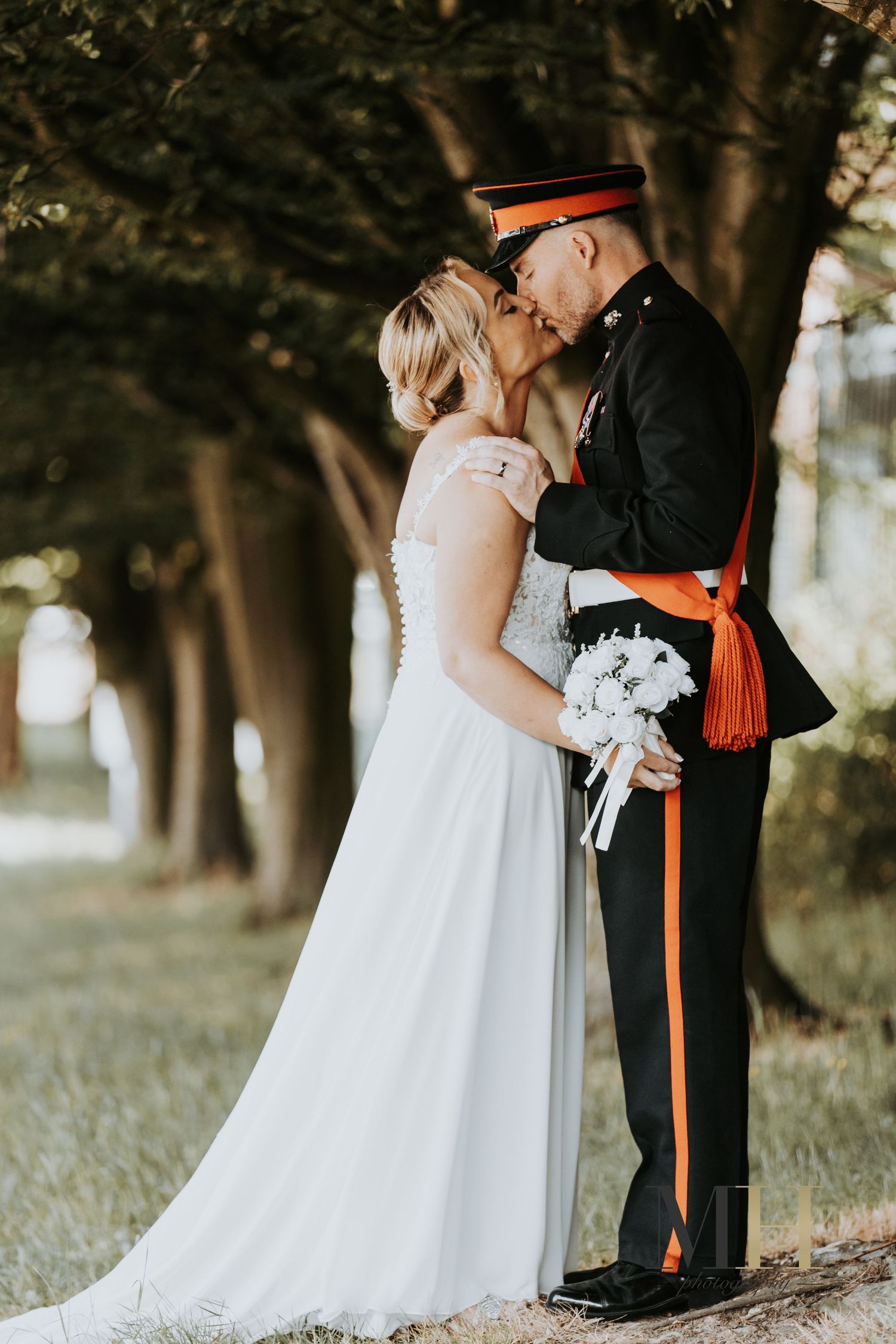 Bride and groom share a romantic kiss outdoors, groom in military dress uniform with orange sash, bride in white gown holding bouquet.