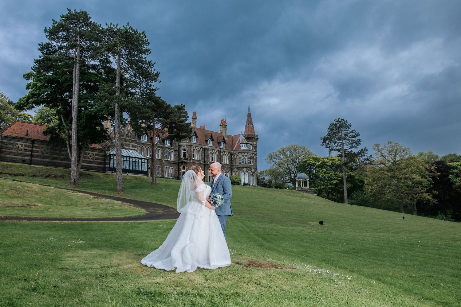 Bride and groom pose on green lawn before grand historic manor under dramatic stormy sky.