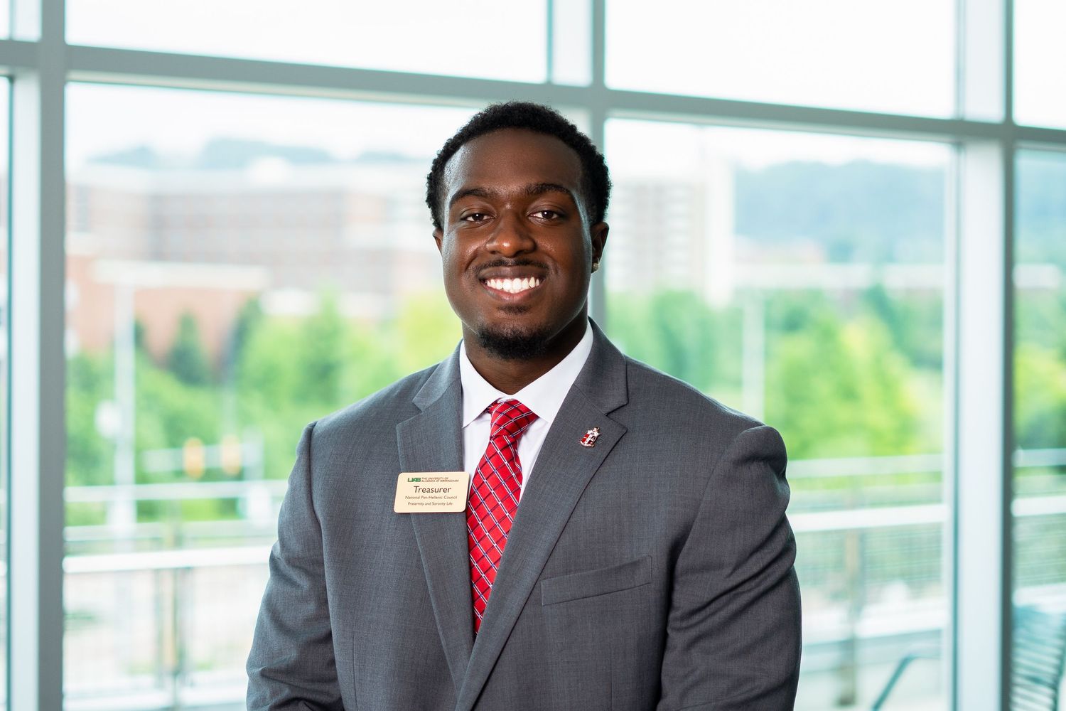 Professional headshot in gray suit and red tie against window background.