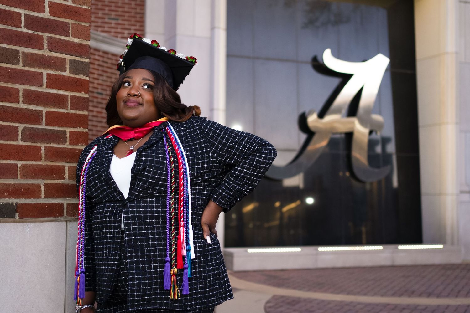Graduate in a black tweed coat with scarf poses confidently against brick wall with storefront window.