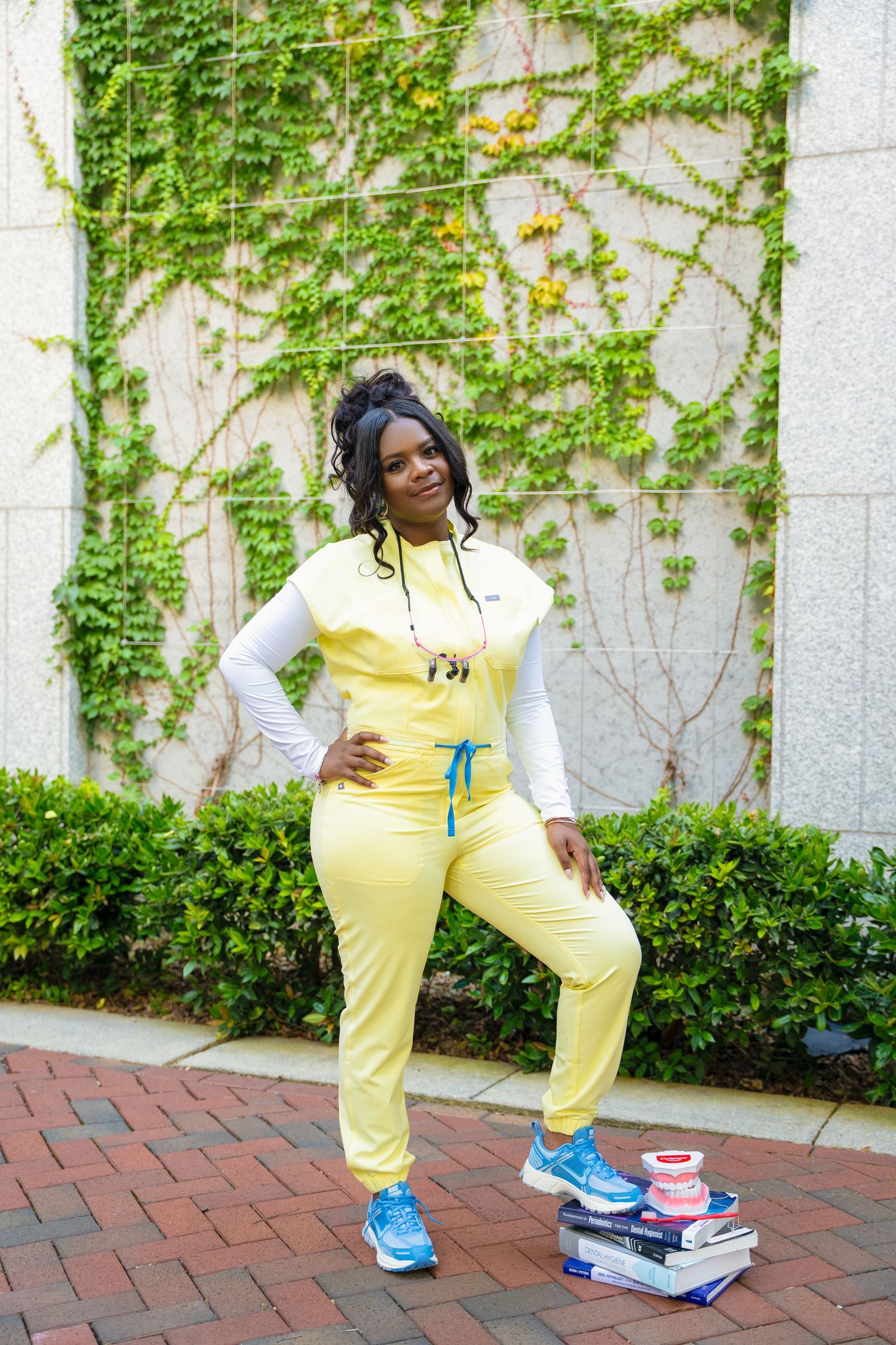 Graduate in yellow jumpsuit poses with books and athletic shoes on brick pathway.