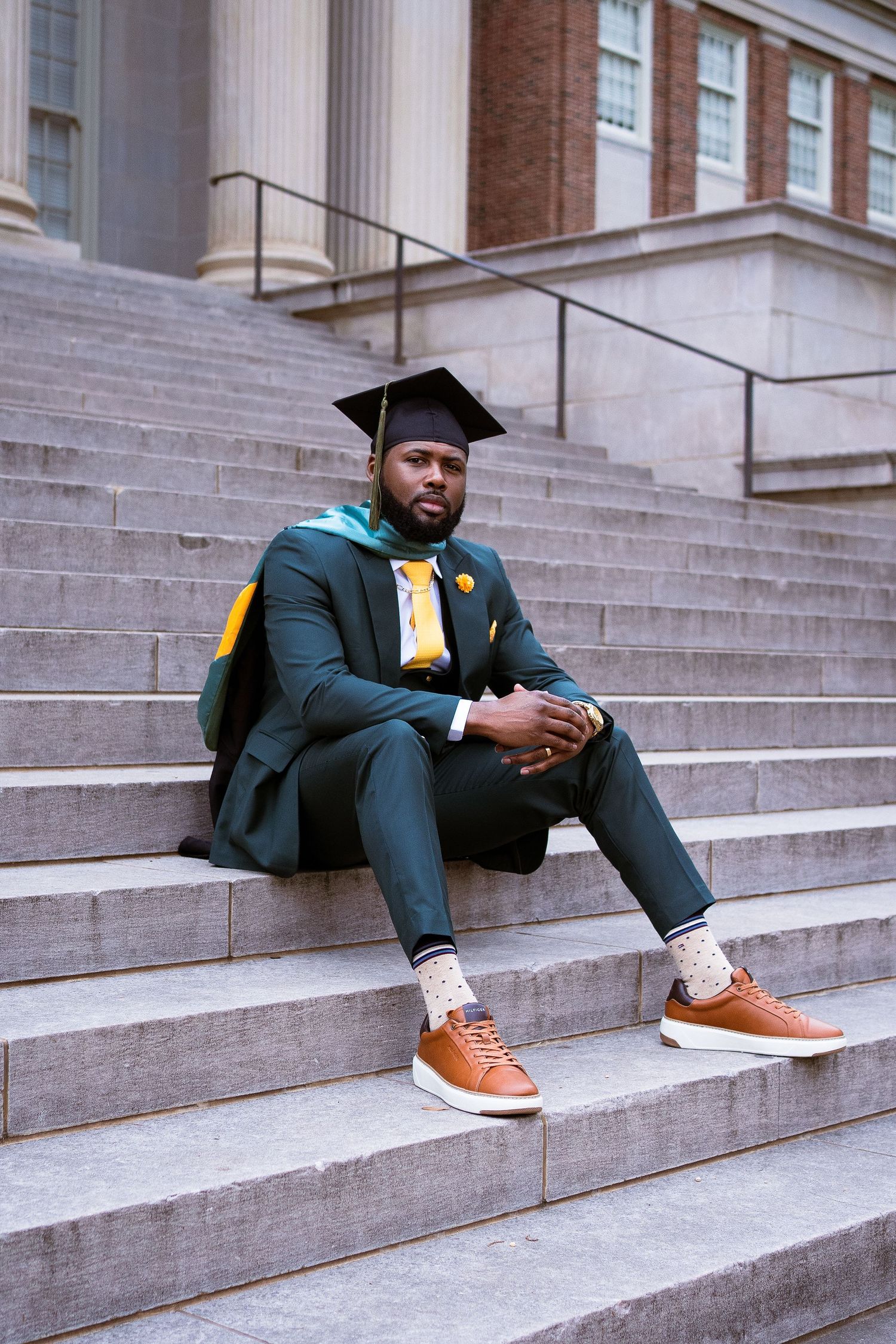 Graduate in green regalia poses on concrete steps of academic building.