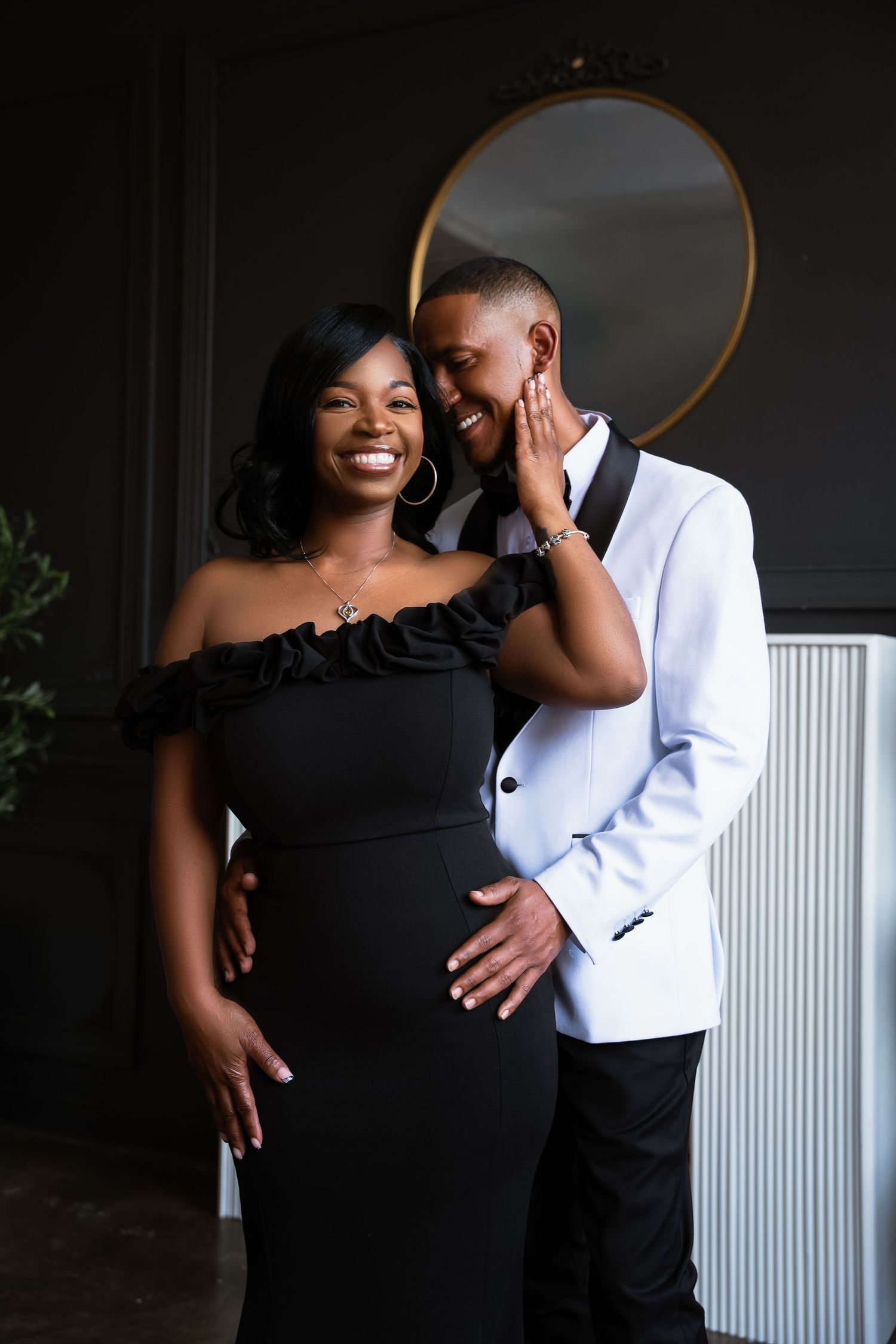 Elegant couple posing together, woman in black off-shoulder gown and man in white tuxedo jacket, sharing a joyful moment.