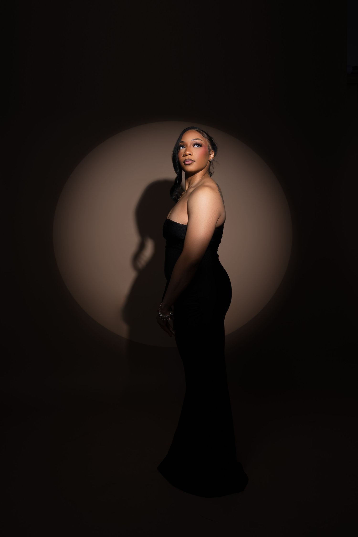 A woman in a strapless black gown poses elegantly against a dark background with a circular spotlight behind her.