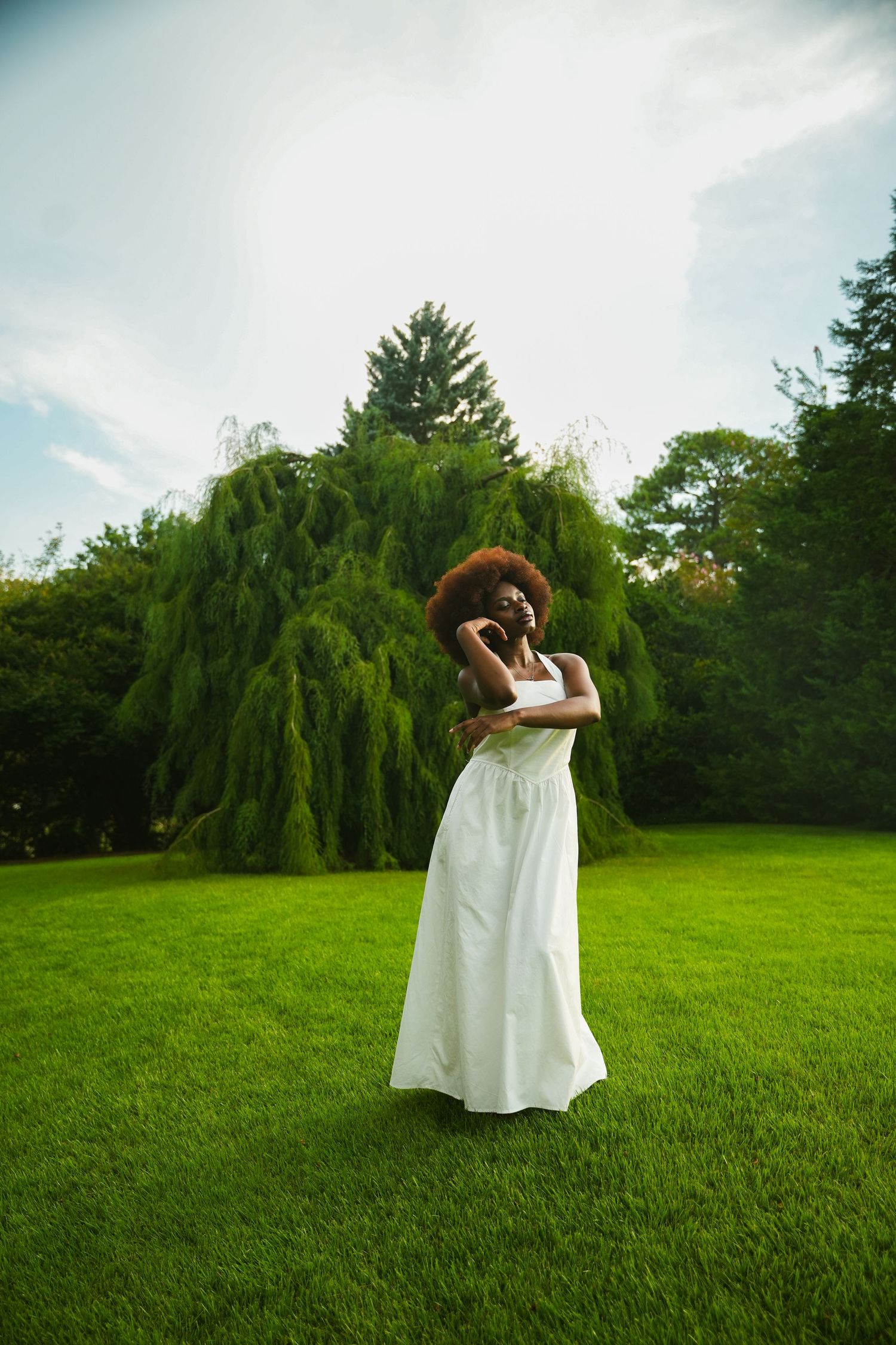 A figure in a flowing white dress poses gracefully on a lush green lawn with weeping willow trees in the background.