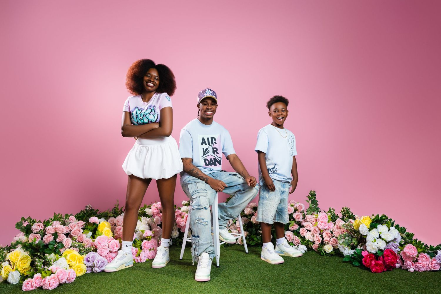 Family members wearing coordinated white outfits pose together against a pink backdrop with colorful flowers.