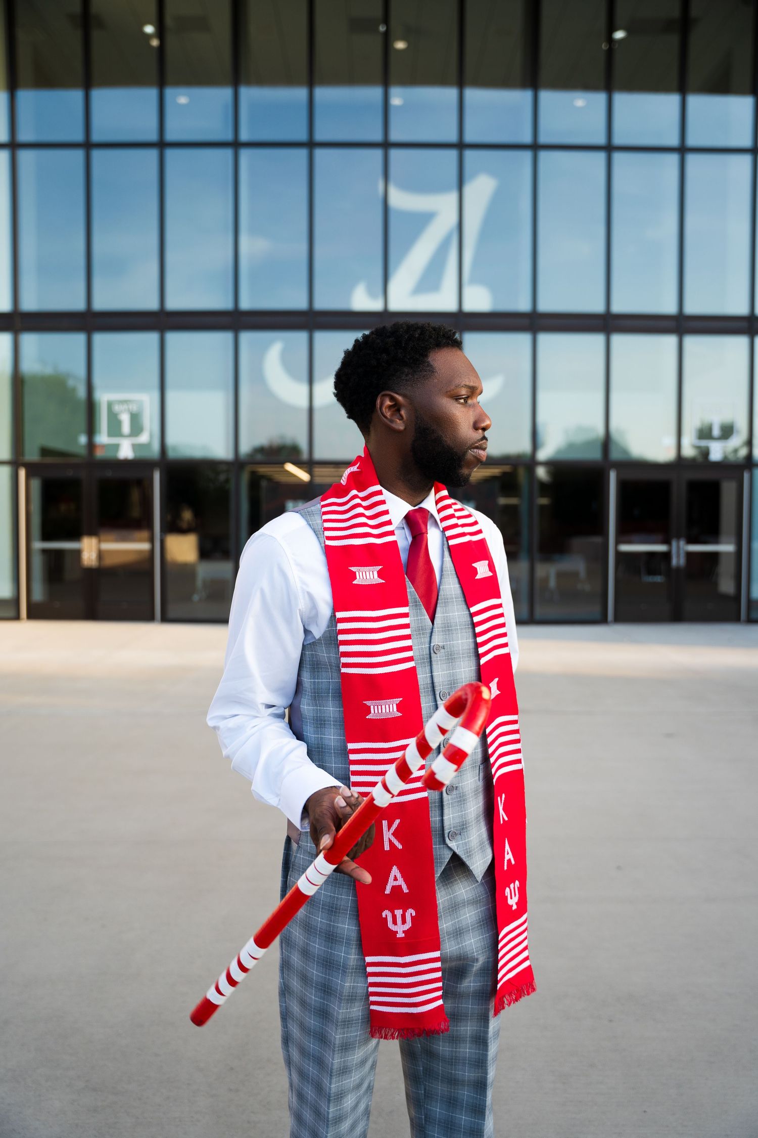 A person wearing a white shirt, plaid suit and red striped Kappa scarf stands in front of a modern building with glass windows.