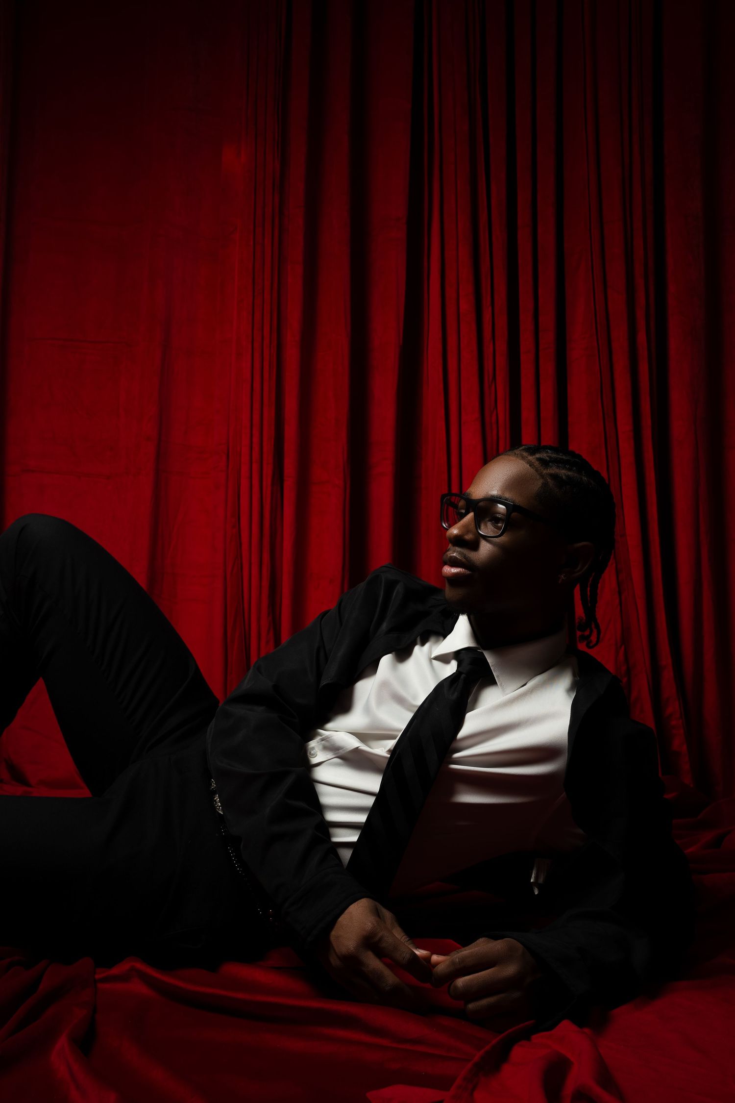 Stylish figure in black suit and white shirt reclines against red curtain backdrop in dramatic low-key lighting.