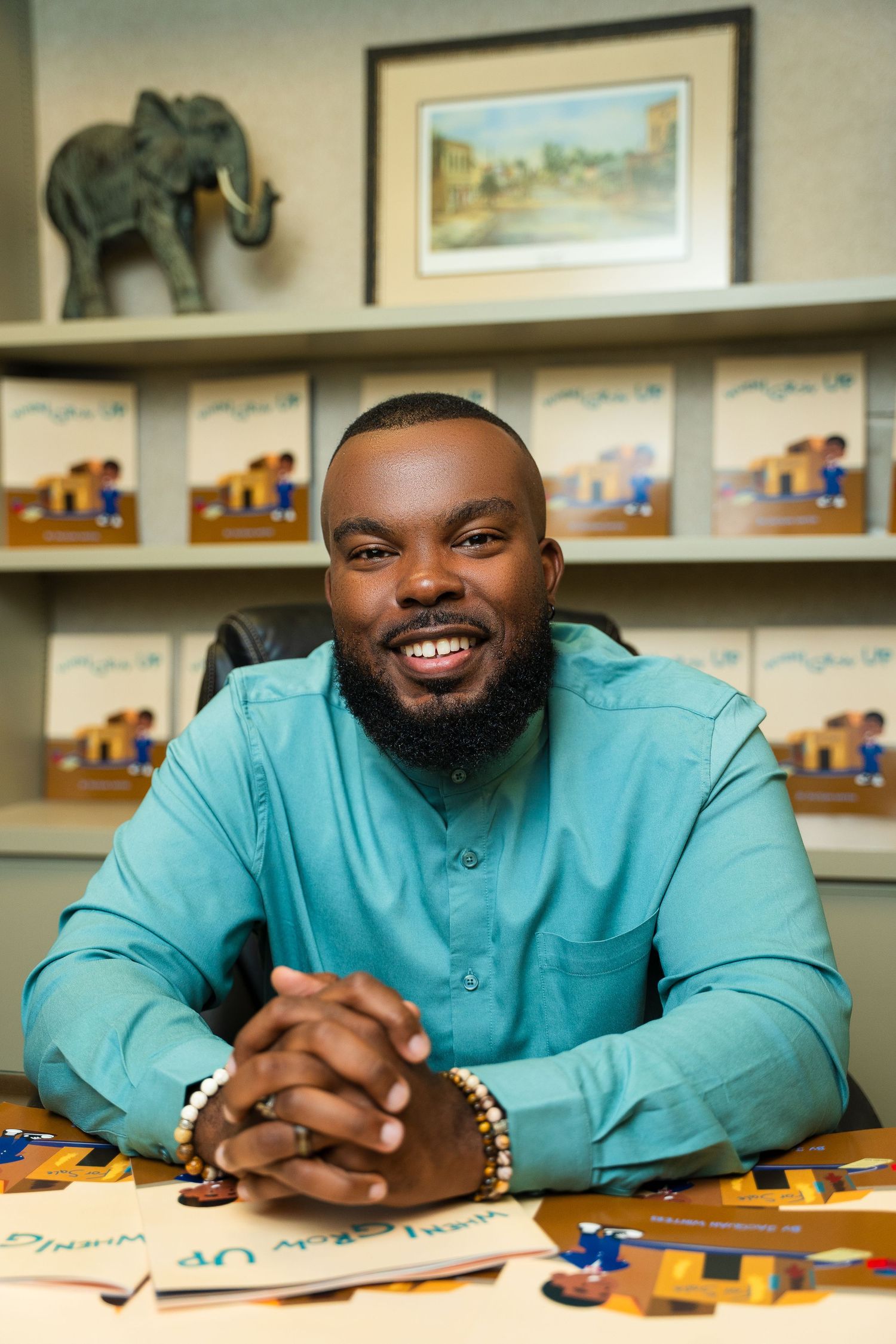 Author seated at desk with children's books and elephant decor in background.