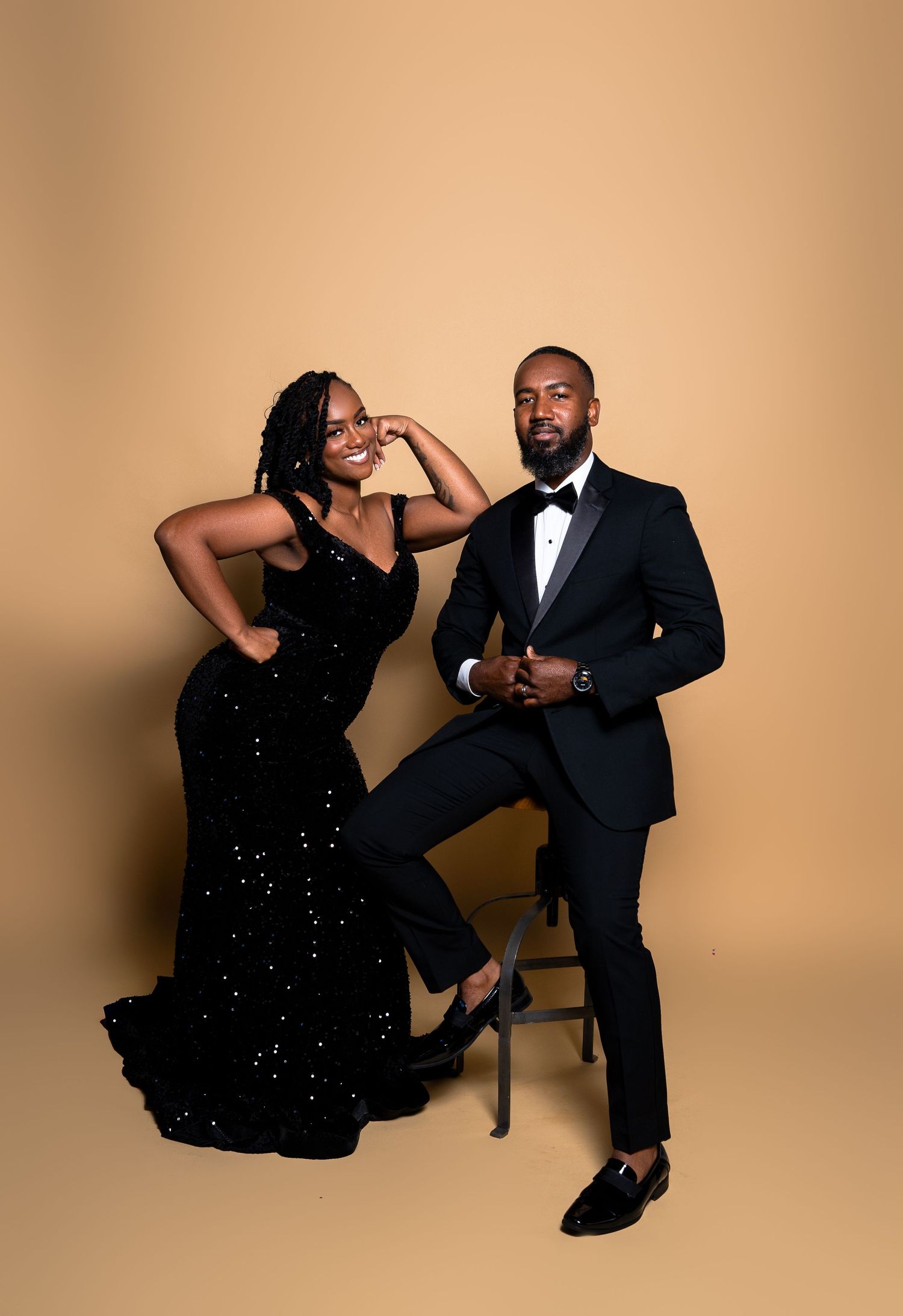 A glamorous posed studio photo of a couple in formal black attire against a beige backdrop.