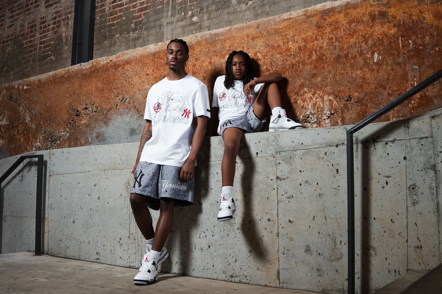People in casual white t-shirts leaning against an industrial brick and concrete wall.
