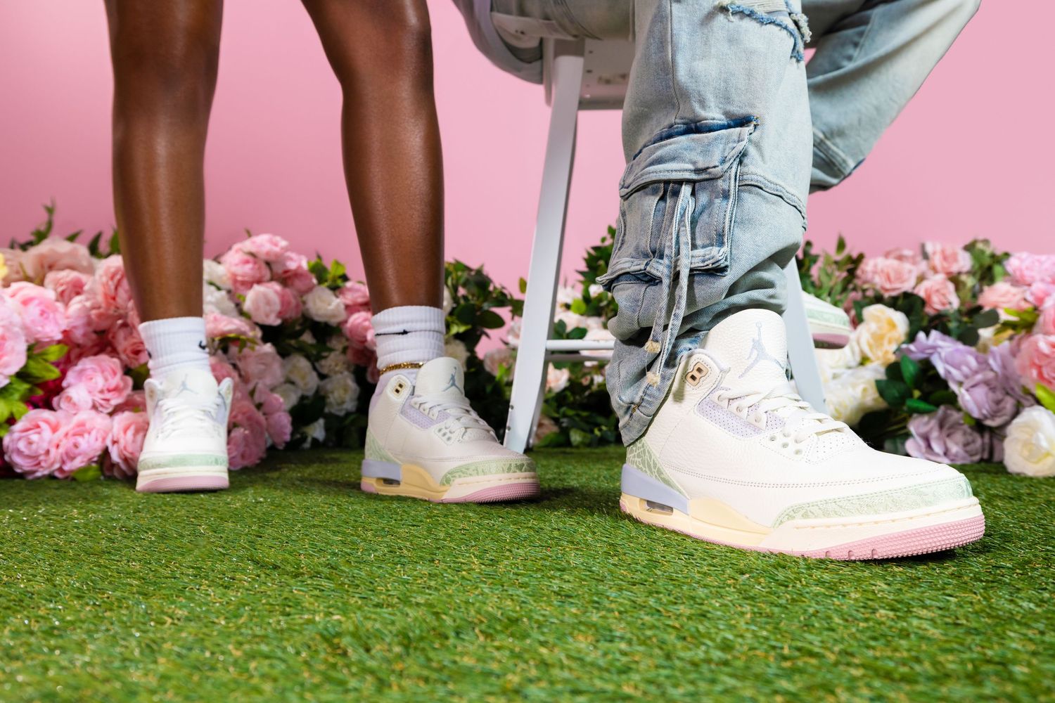 Two pairs of white sneakers on artificial grass with pink flowers in the background against a pink wall.
